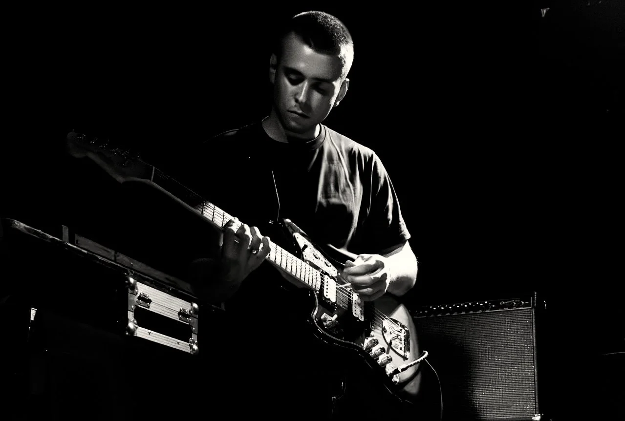 A young man playing an electric guitar on stage, black background, in black and white.