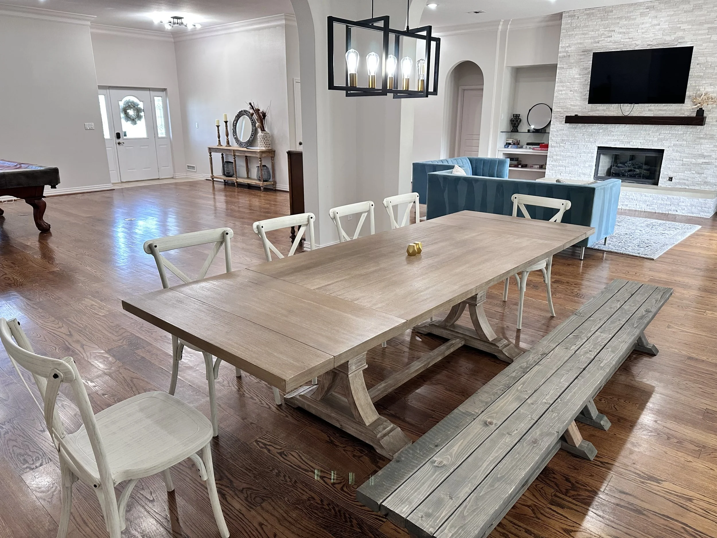 Open living area with dining table, chairs, and bench, wooden flooring, modern chandelier, fireplace, wall-mounted TV, and blue sofas.