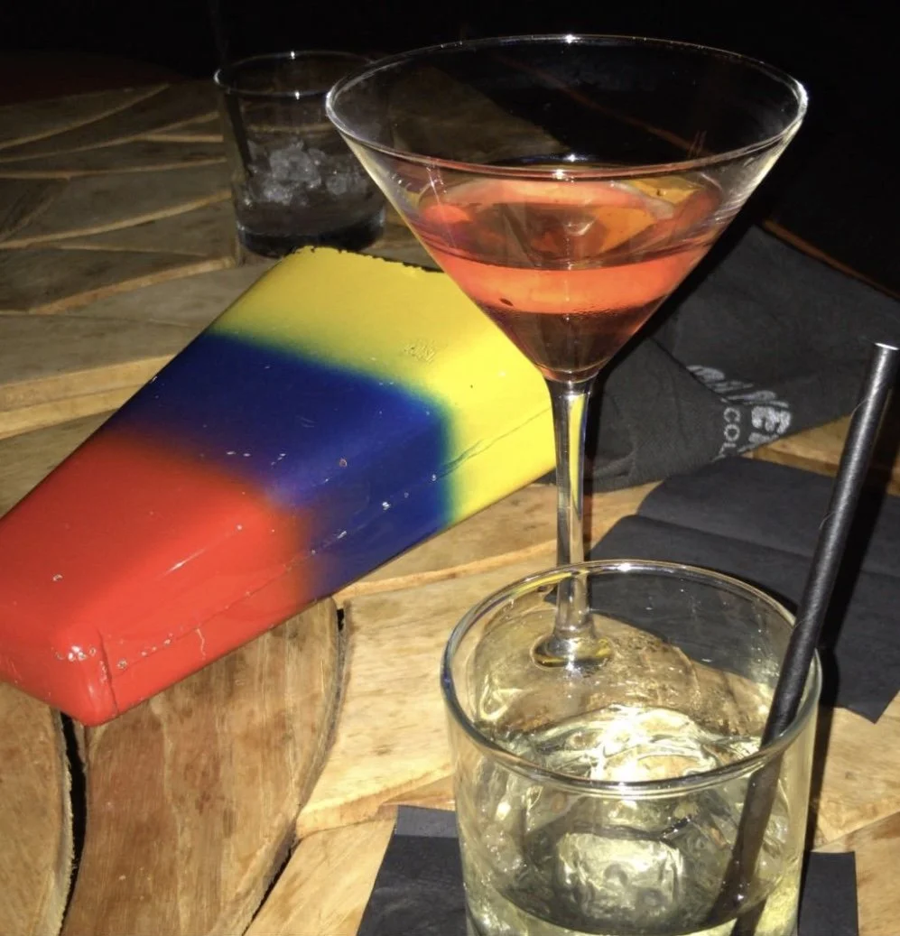 Colorful coffin prop on table with cocktail glasses, one with pink drink, on dark napkins.