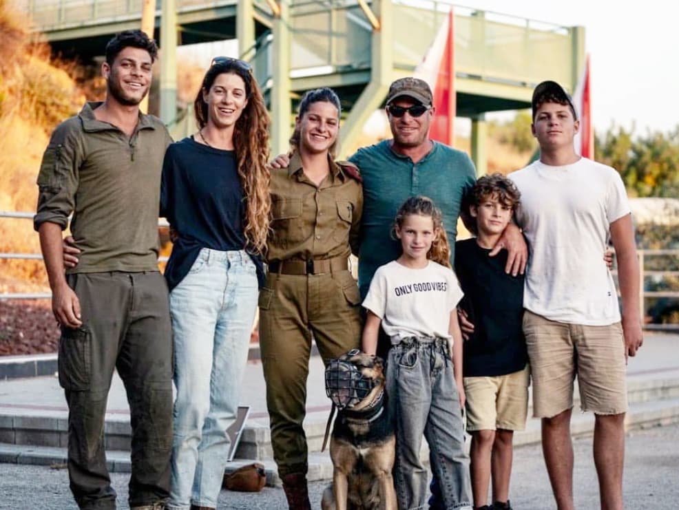 Group of seven people, including a woman in military uniform, standing outdoors in front of a slide structure, smiling for a photo, with a dog.