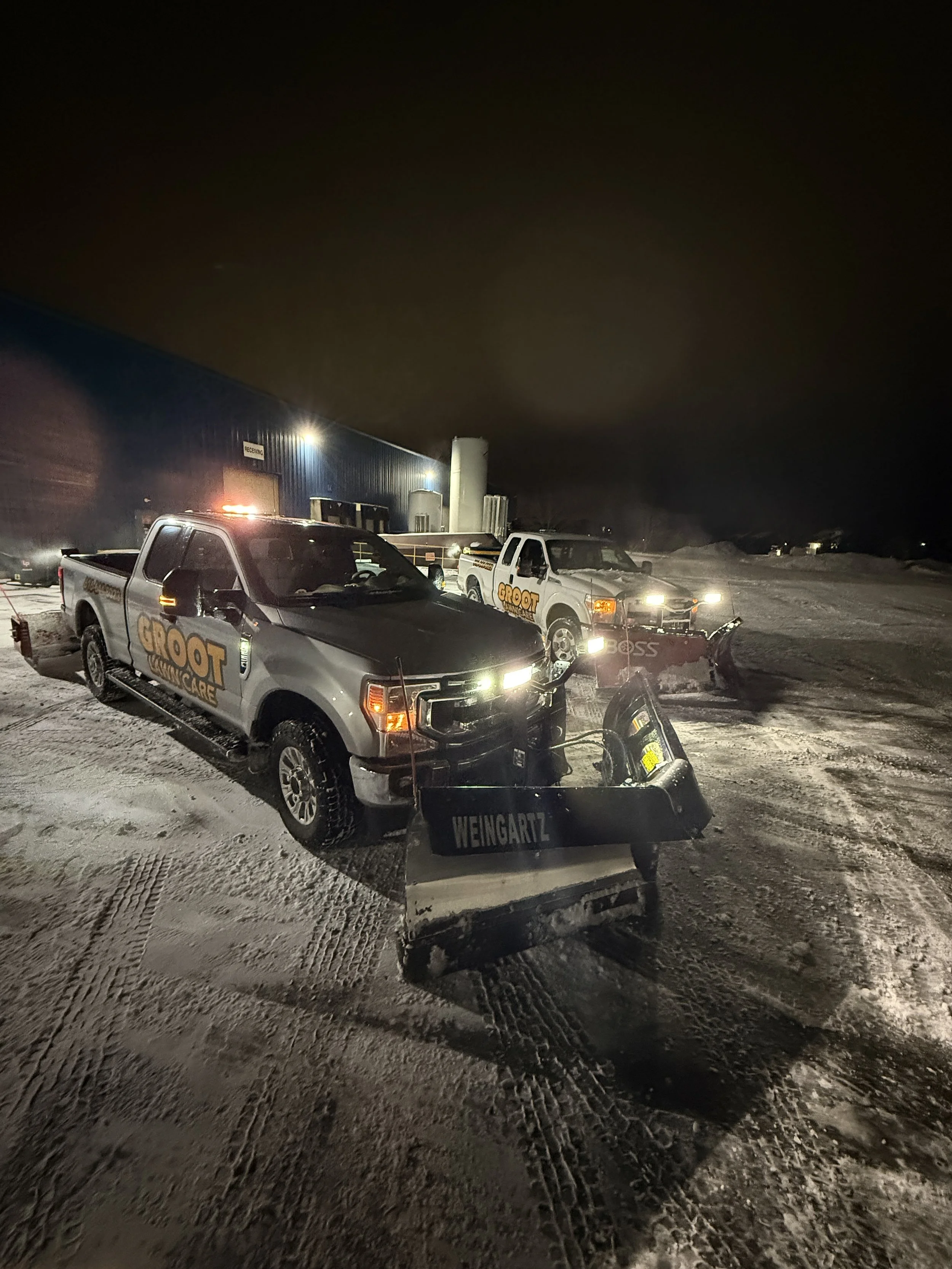 Nighttime scene with two pickup trucks and snow removal equipment, branded with 'Groot' and 'Weingartz', parked on a snow-covered lot outside a large industrial building.