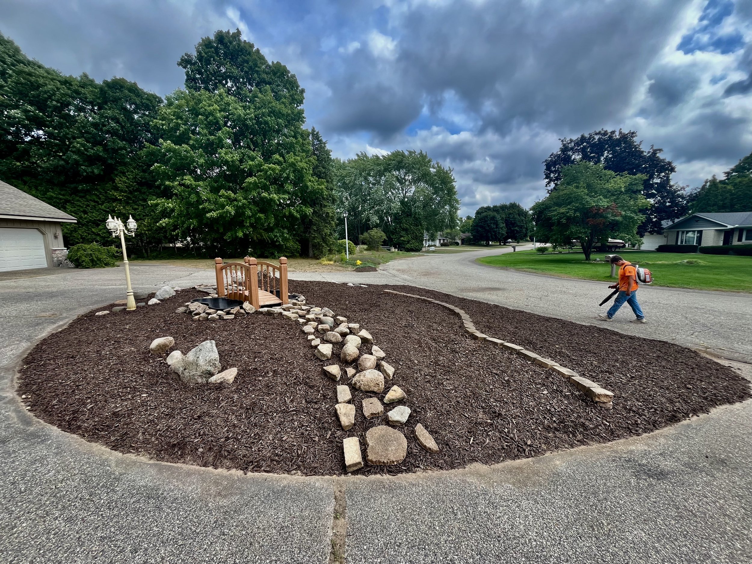 A landscaped roundabout with dark mulch, a small wooden bridge, and a line of rocks on a curved path, with a man in an orange shirt trimming the mulch with a leaf blower, suburban houses, trees, and a cloudy sky in the background.