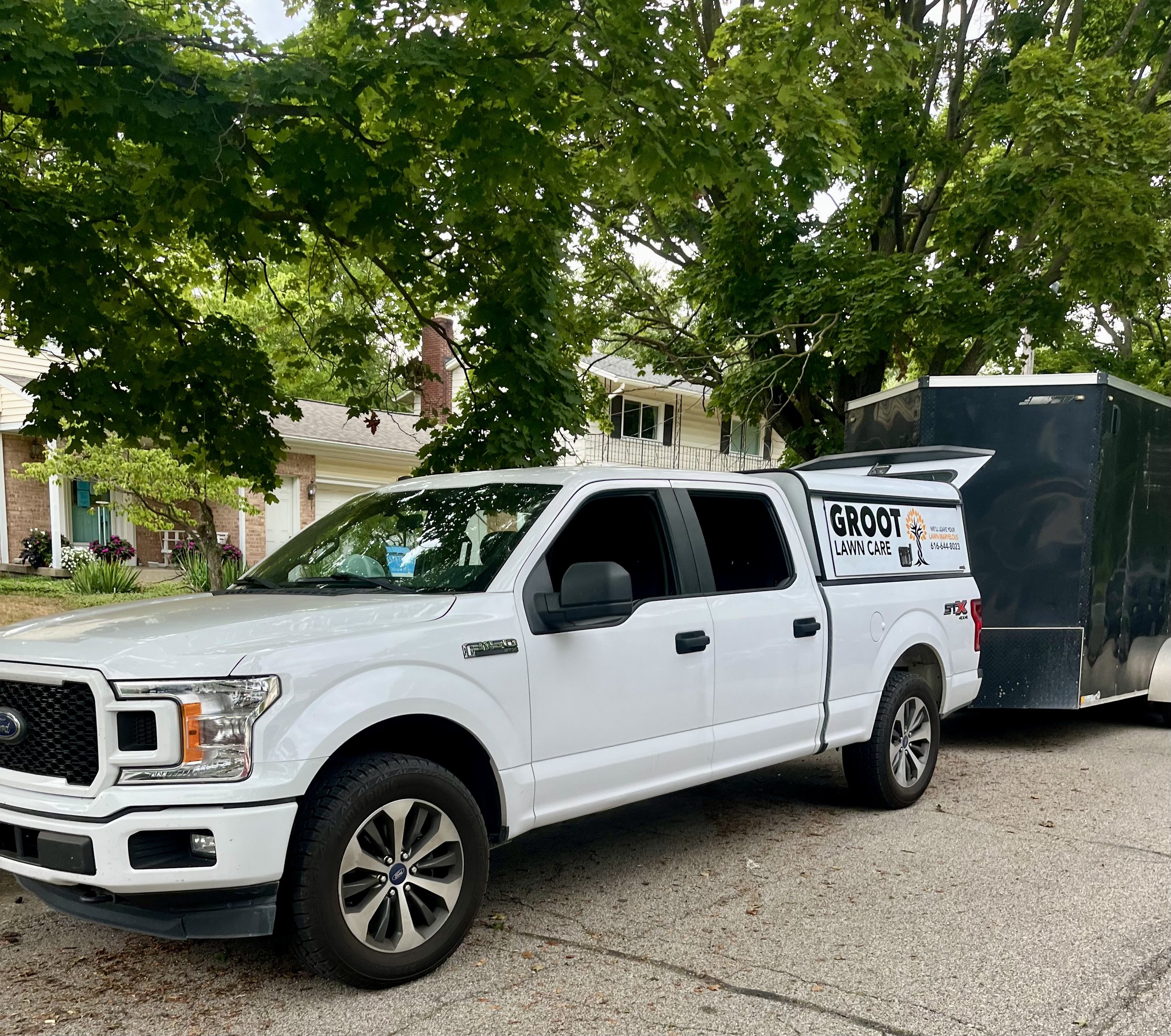 A white Ford F-150 pickup truck attached to a black trailer, parked on a suburban street under large leafy tree. The truck has a sign on the side that reads "GROOT LAWN CARE" with contact information.