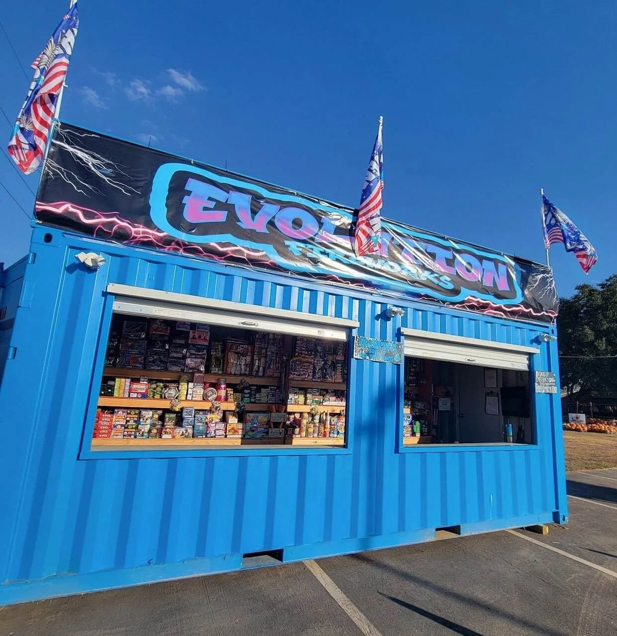 Blue shipping container stand selling fireworks near me and fireworks in Katy, TX, with American flags and an EVOLUTION sign on top against a clear sky.