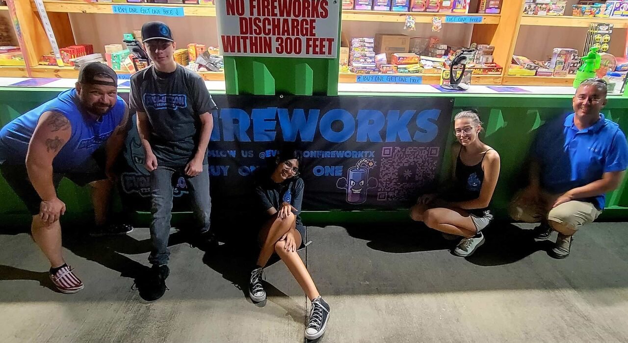 Five people pose at a fireworks near me stand at night; shelves of fireworks and a “No Fireworks Discharge Within 300 Feet” sign are visible.
