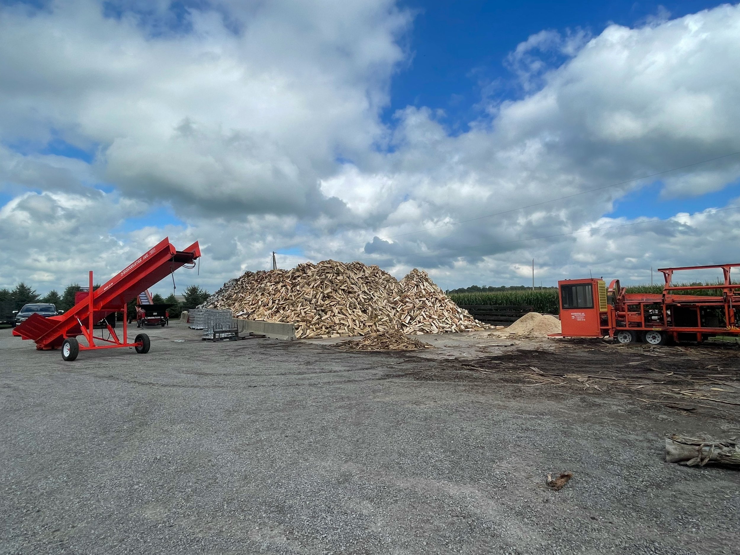 Large pile of cut firewood with red machinery and equipment in an outdoor setting, surrounded by fields under a cloudy sky.