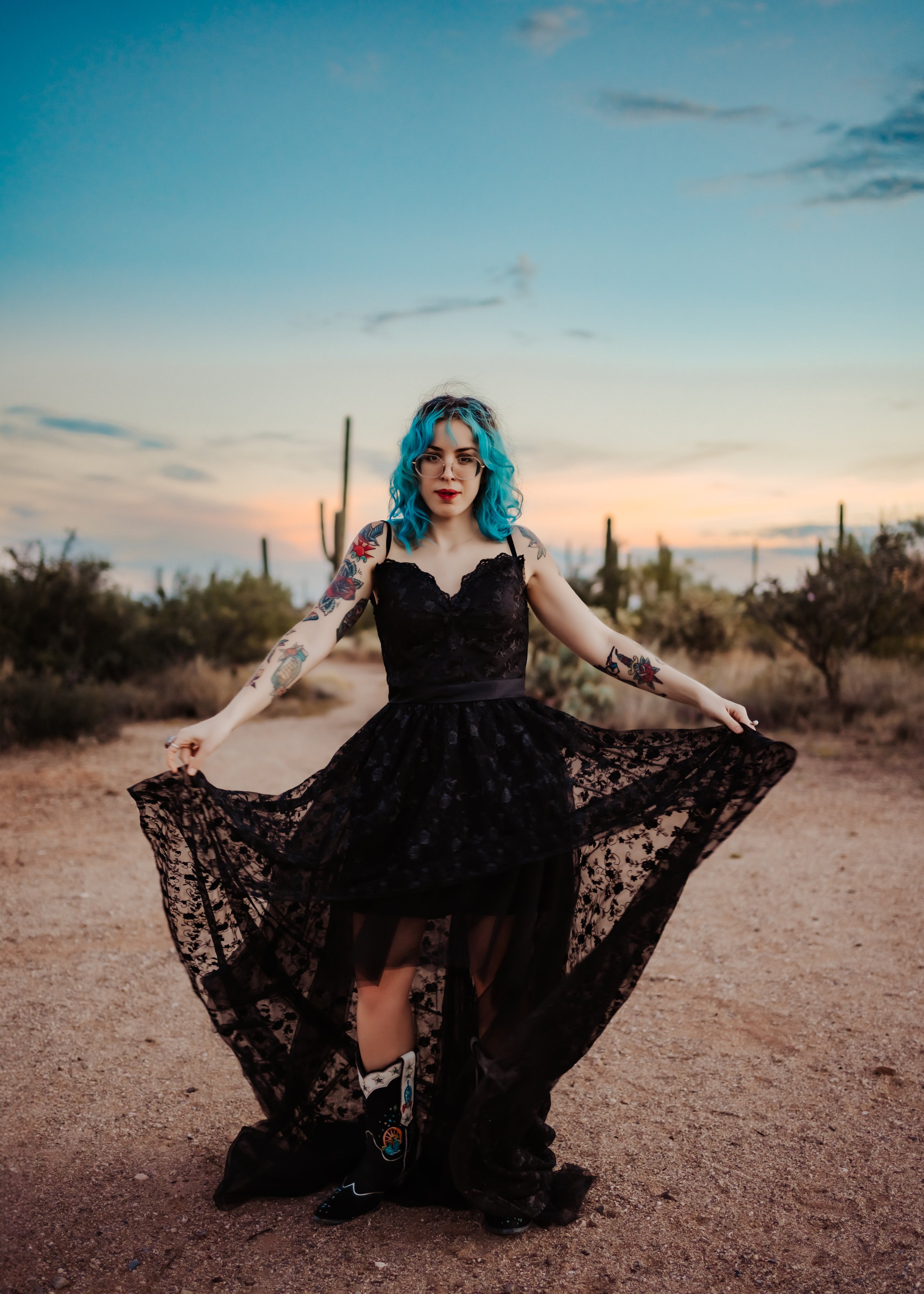 A woman with blue hair in a black lace dress and cowboy boots poses in a desert landscape with cacti at sunset.