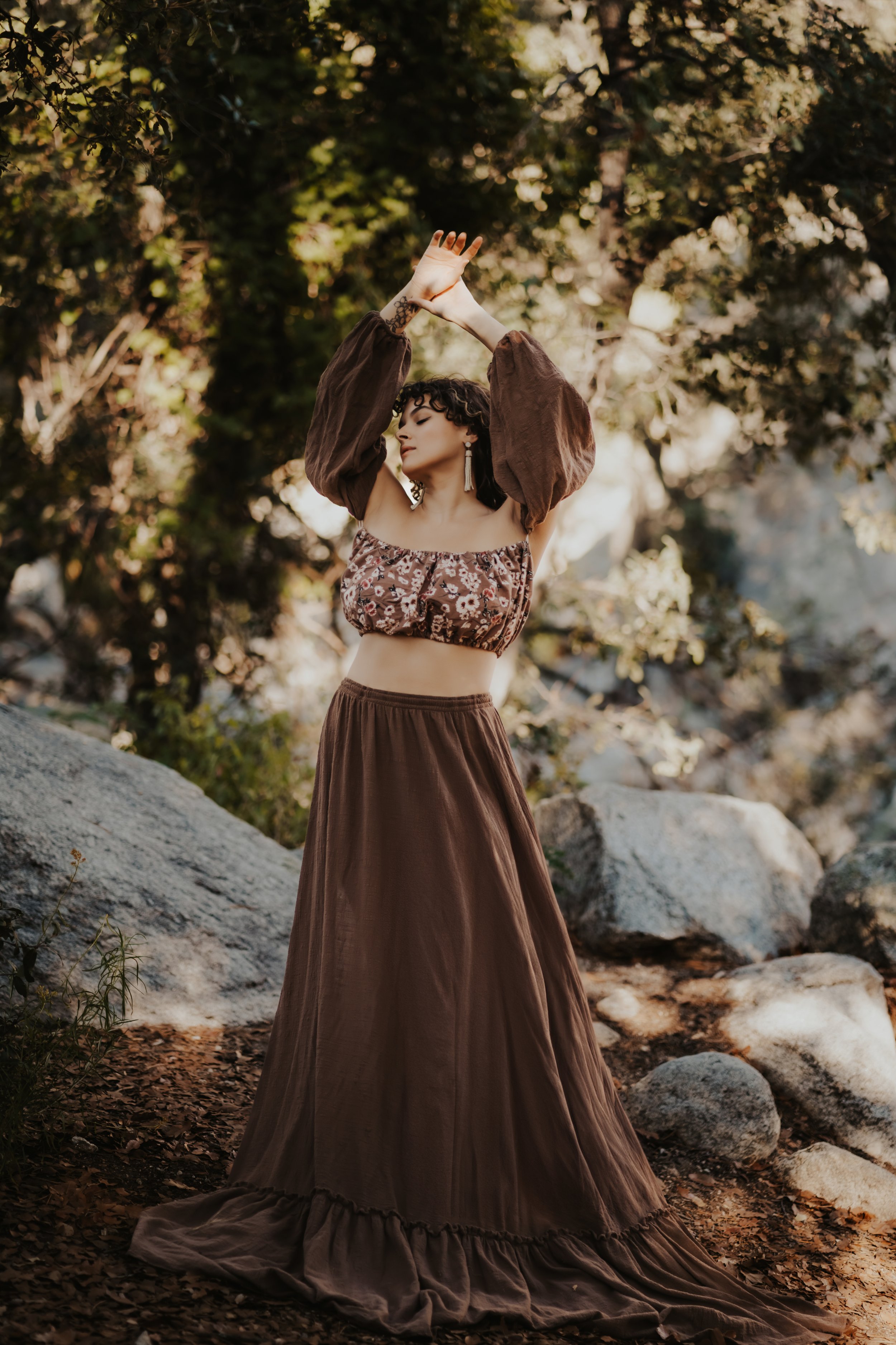 Woman in brown floral top and long skirt poses with arms raised in a forest setting.