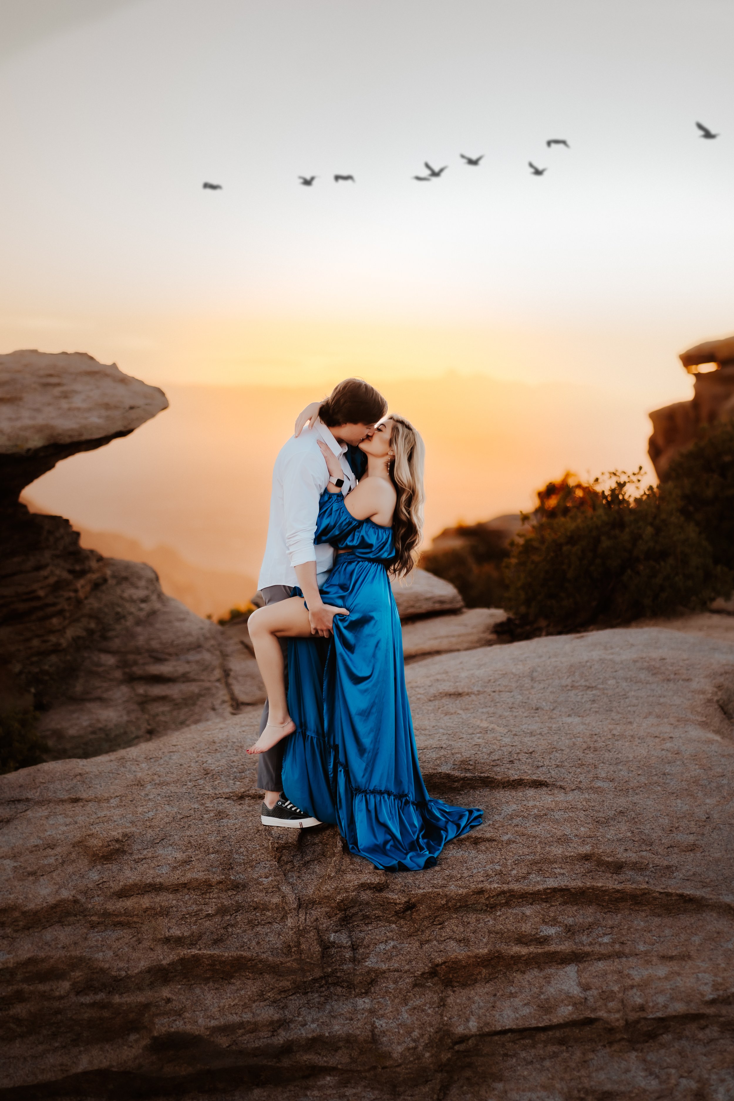 A couple embraces on a rocky cliff at sunset with a flock of birds flying above. The woman wears a blue dress and the man is in casual attire.