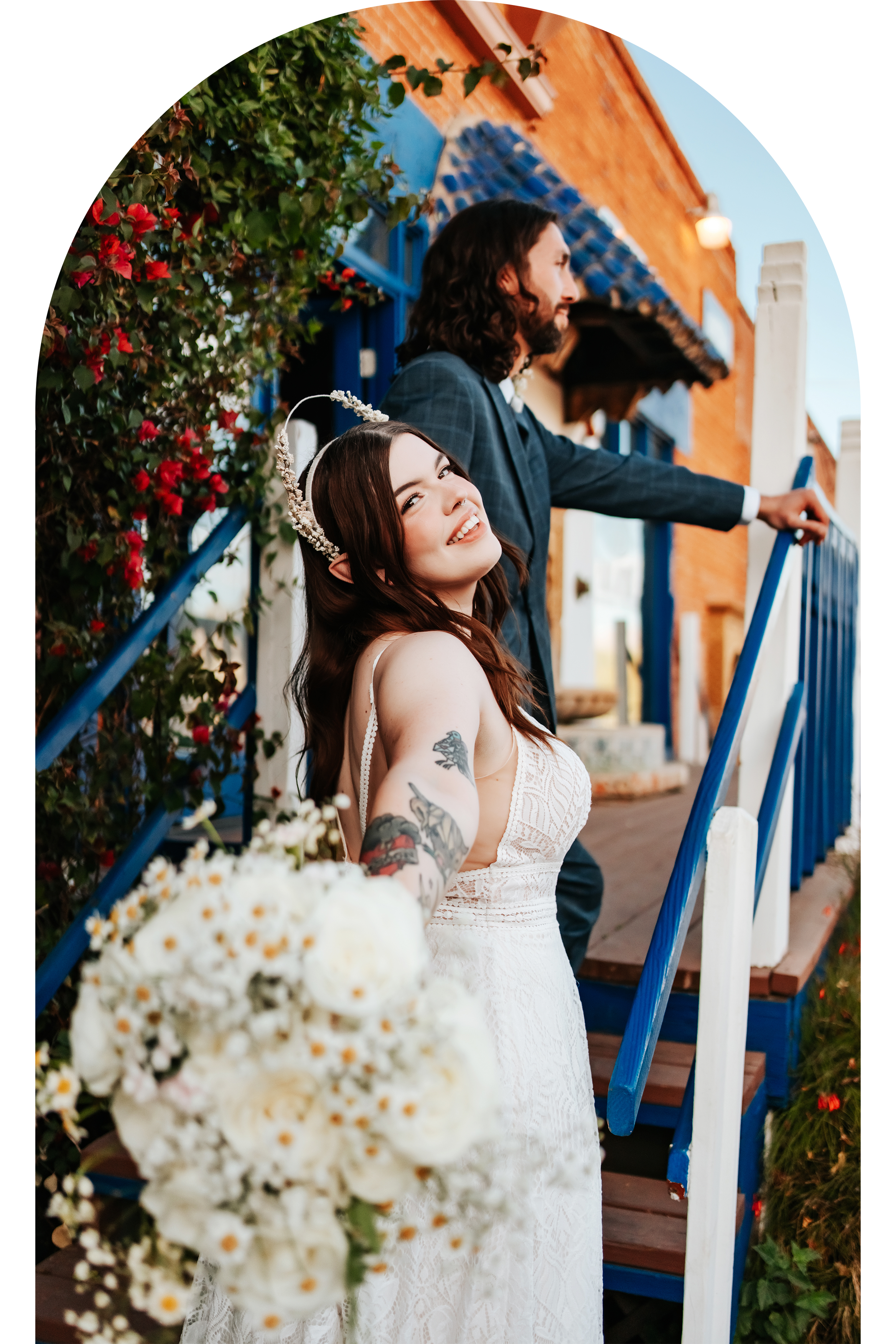 Bride holding a bouquet and smiling, with groom standing on stairs next to colorful building and flowers.