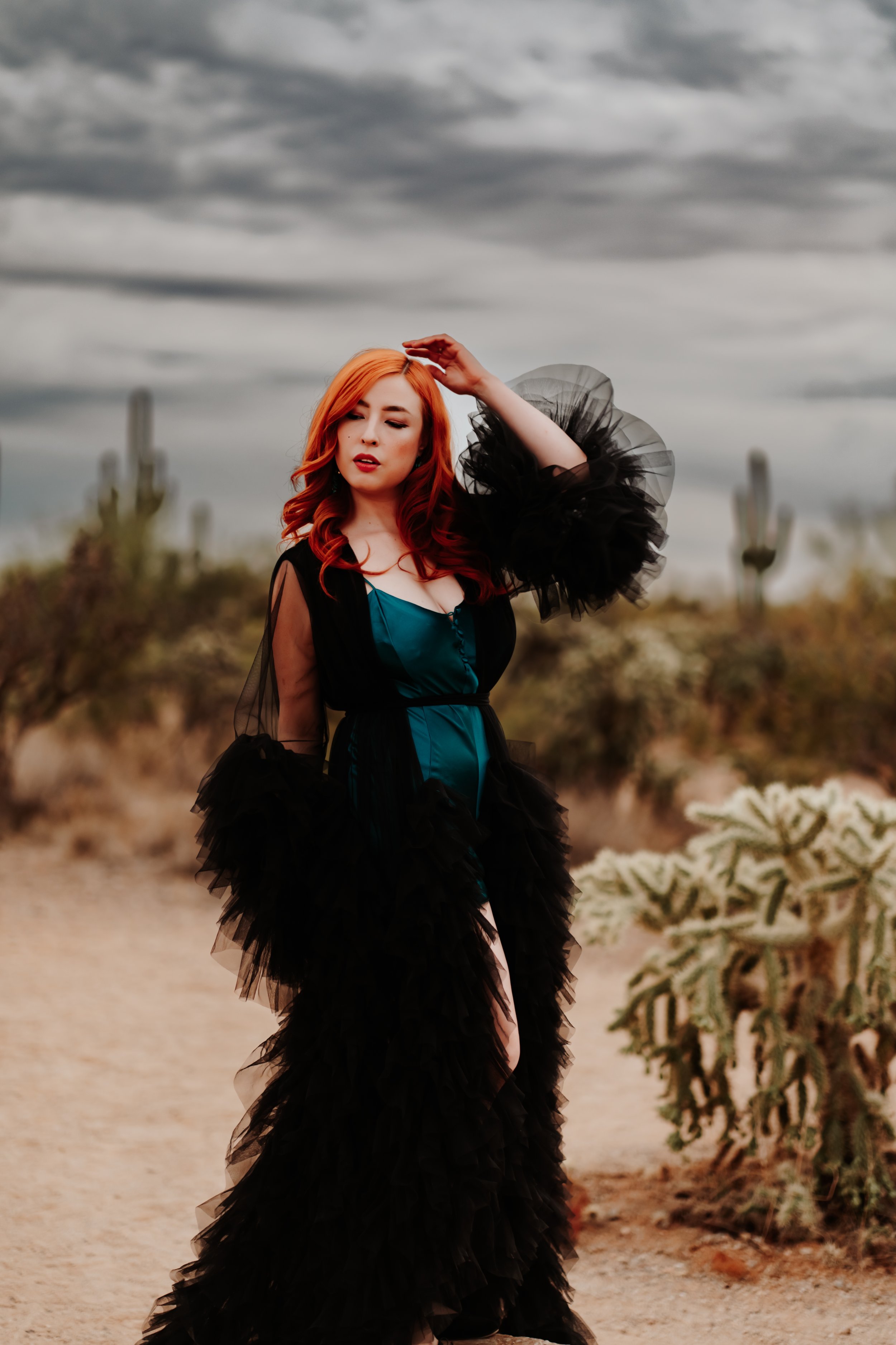 Woman with red hair in black and teal outfit standing in desert landscape with cacti and cloudy sky.