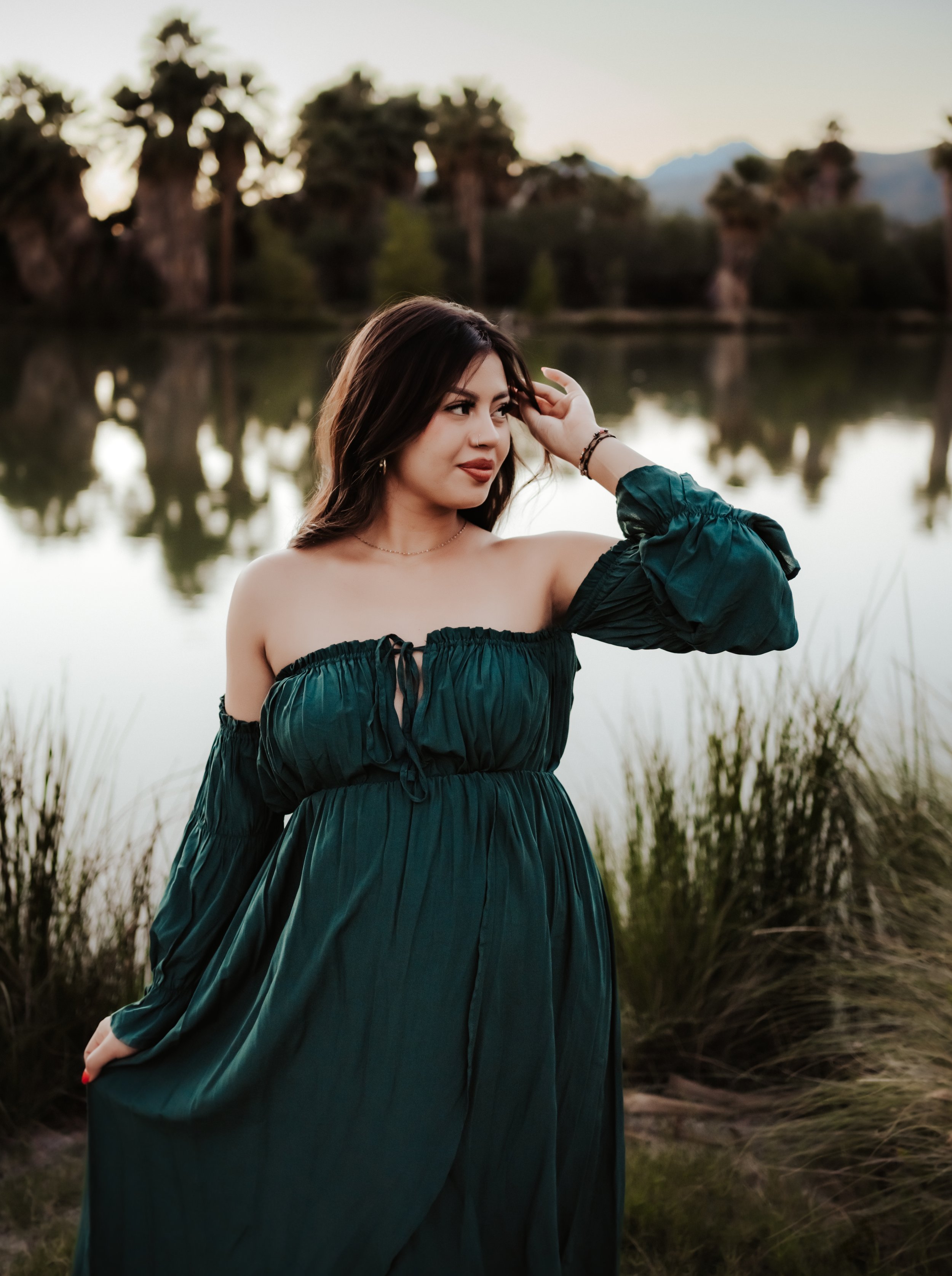 Woman in green off-the-shoulder dress by a lake with palm trees in the background.
