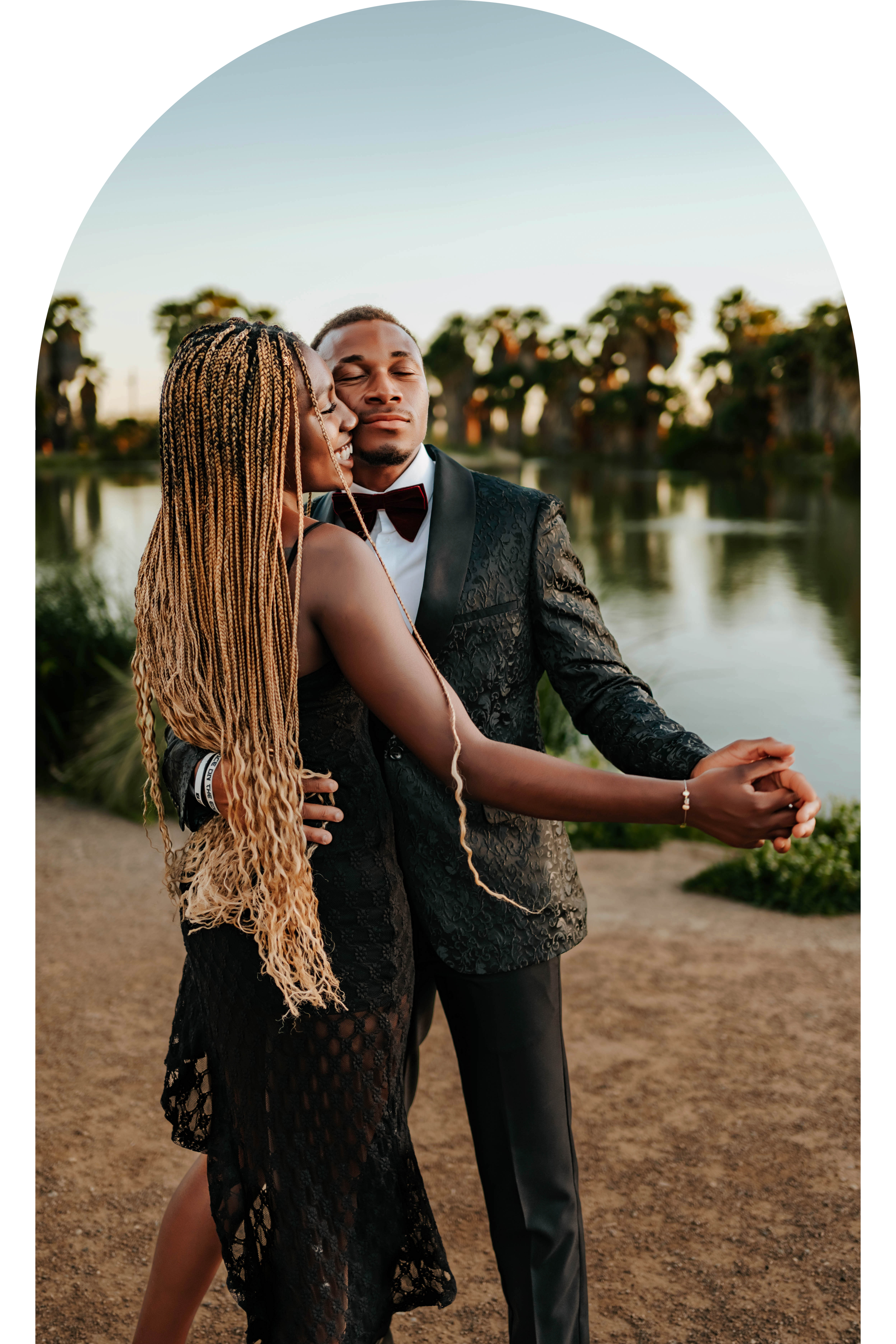 Couple dancing outdoors near a lake, wearing formal attire; woman in a black dress, man in a tuxedo with a bow tie.