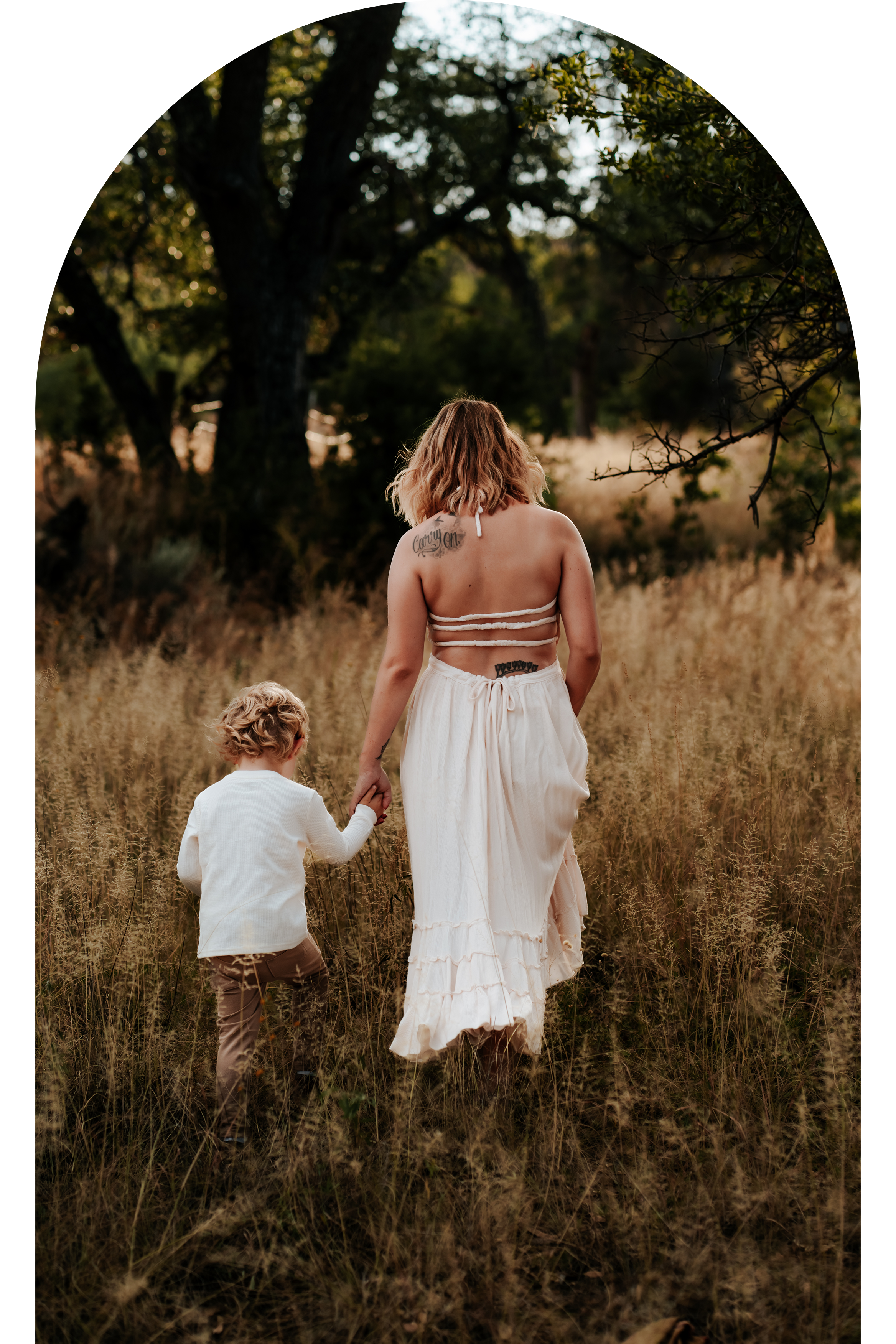 Woman in white dress holding hands with a child, walking through a grassy field with trees in the background.