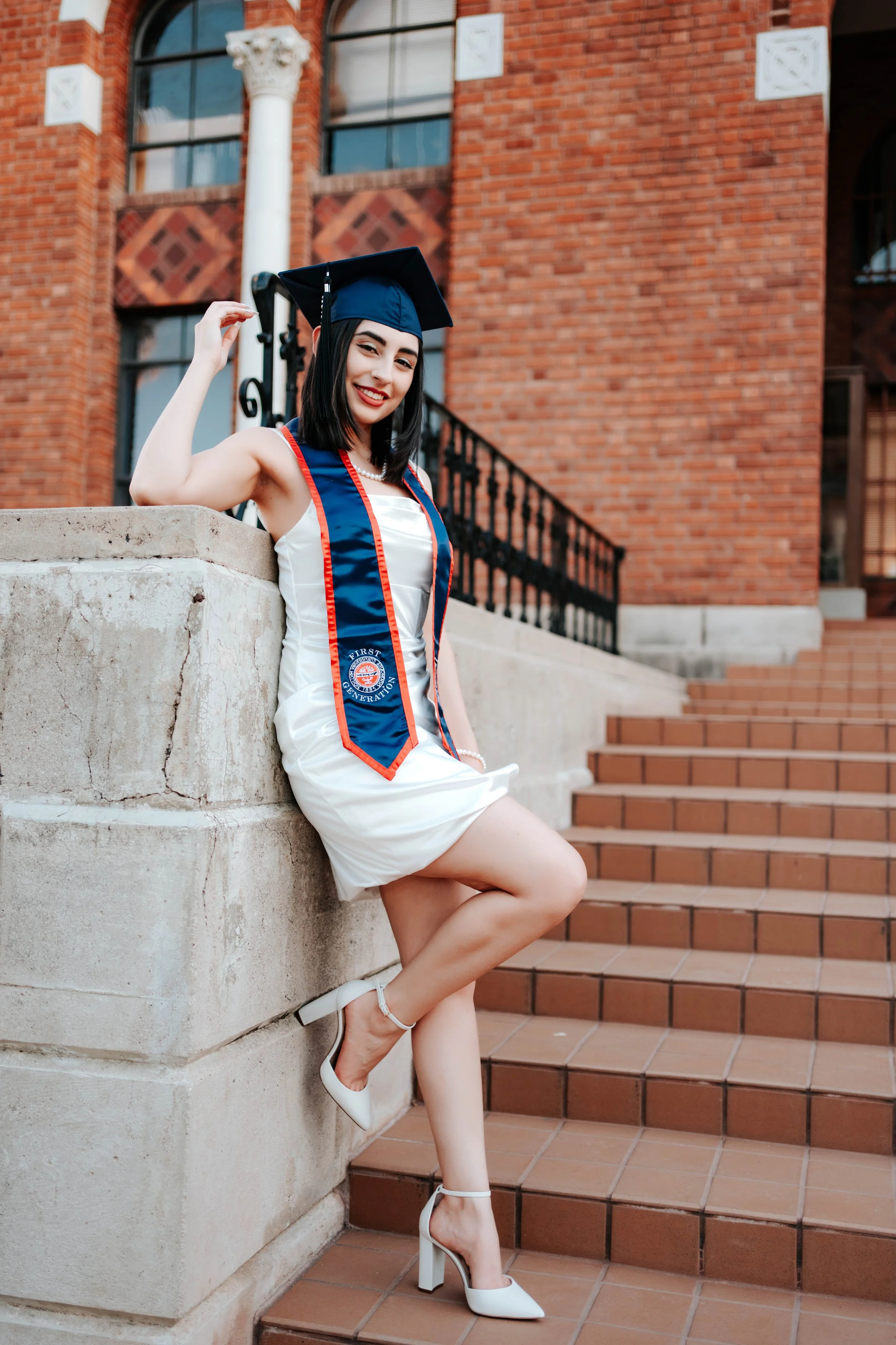 A woman graduating smiles at the camera in front of an academic building on the University of Arizona campus for a senior graduate photoshoot.