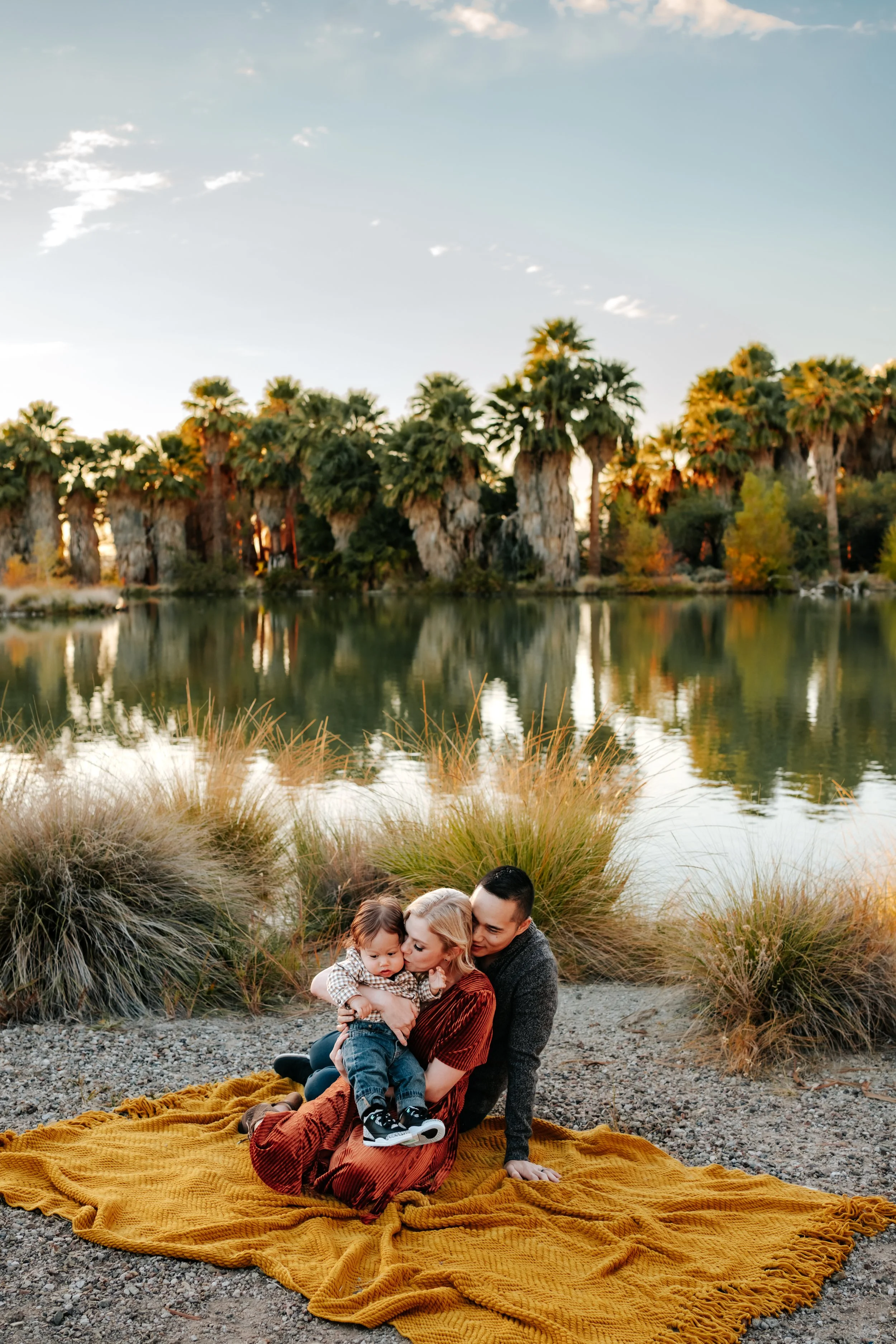 Family sitting on a yellow blanket by a lake, surrounded by tall grass and palm trees, with a clear blue sky.