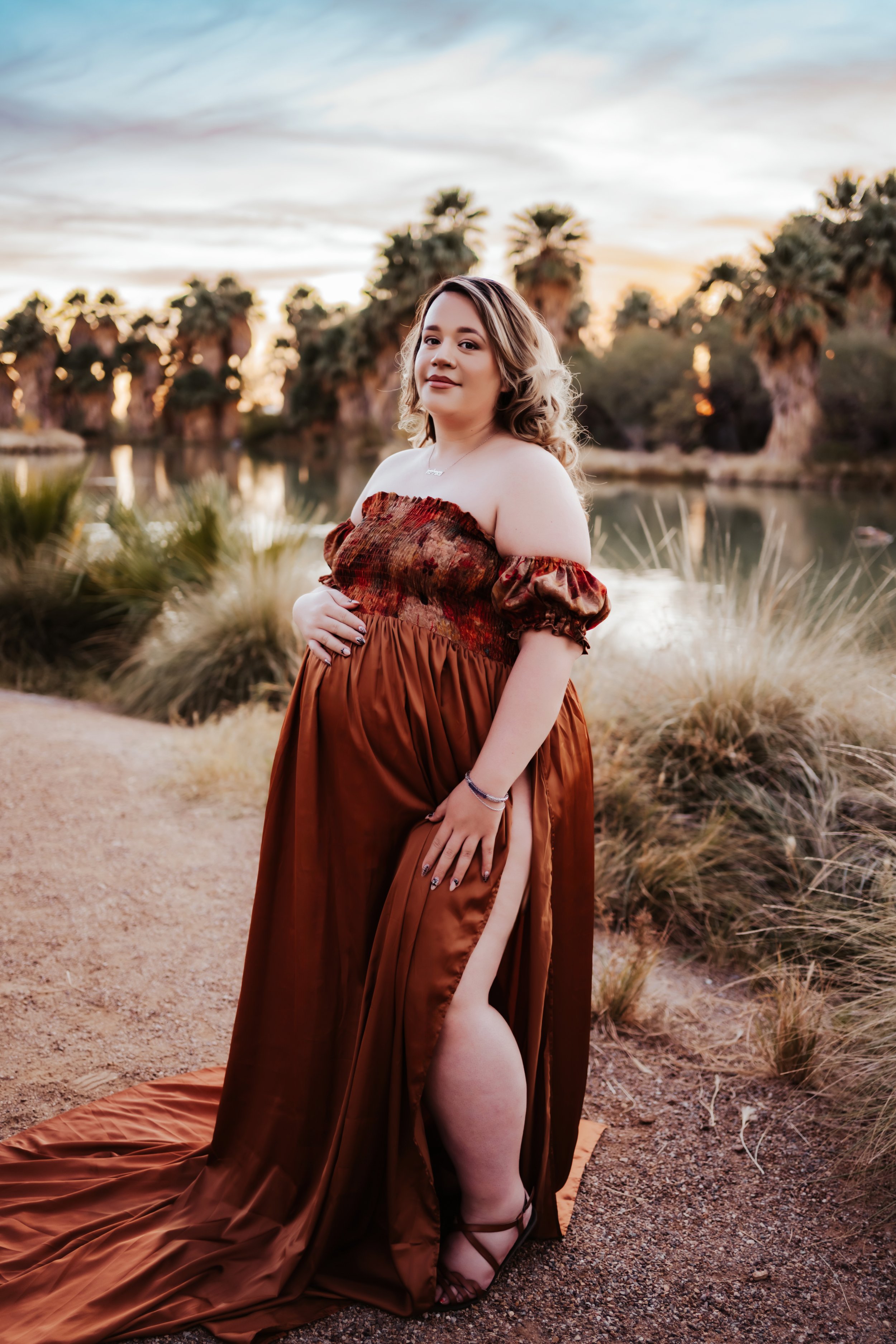 Pregnant woman in brown gown poses outdoors near palm trees.