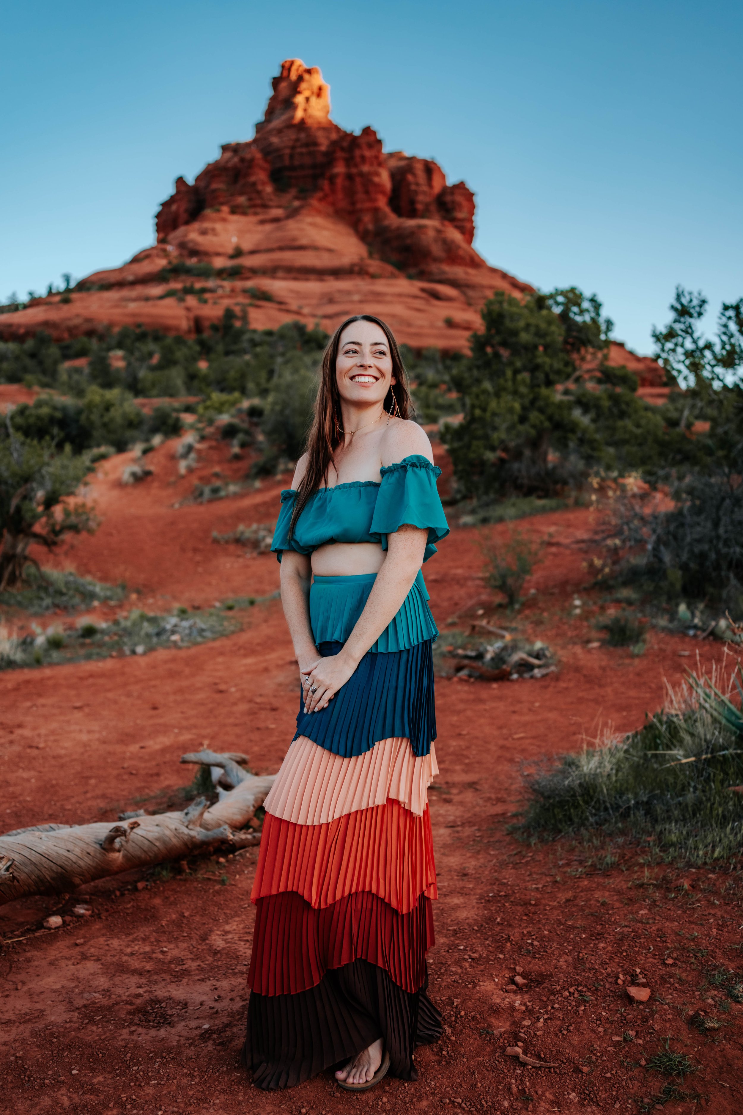 Woman smiling in colorful dress standing in a desert landscape with red rock formations and greenery.