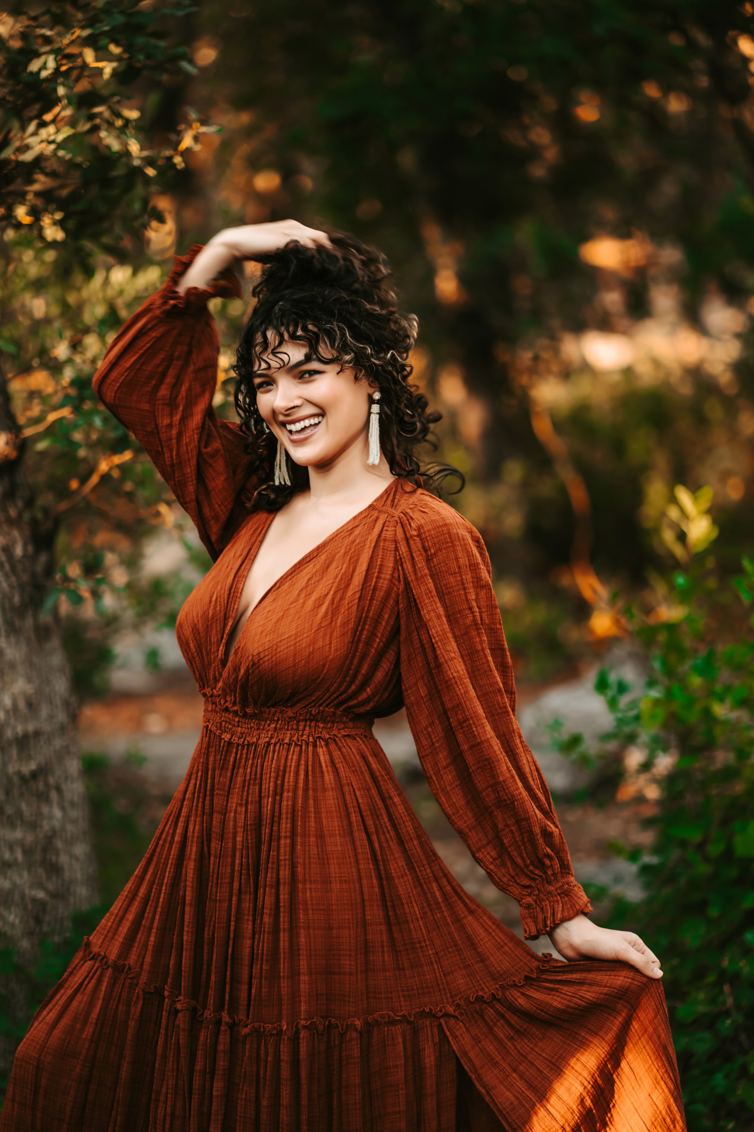 Smiling woman in a long, rust-colored dress poses outdoors surrounded by greenery.