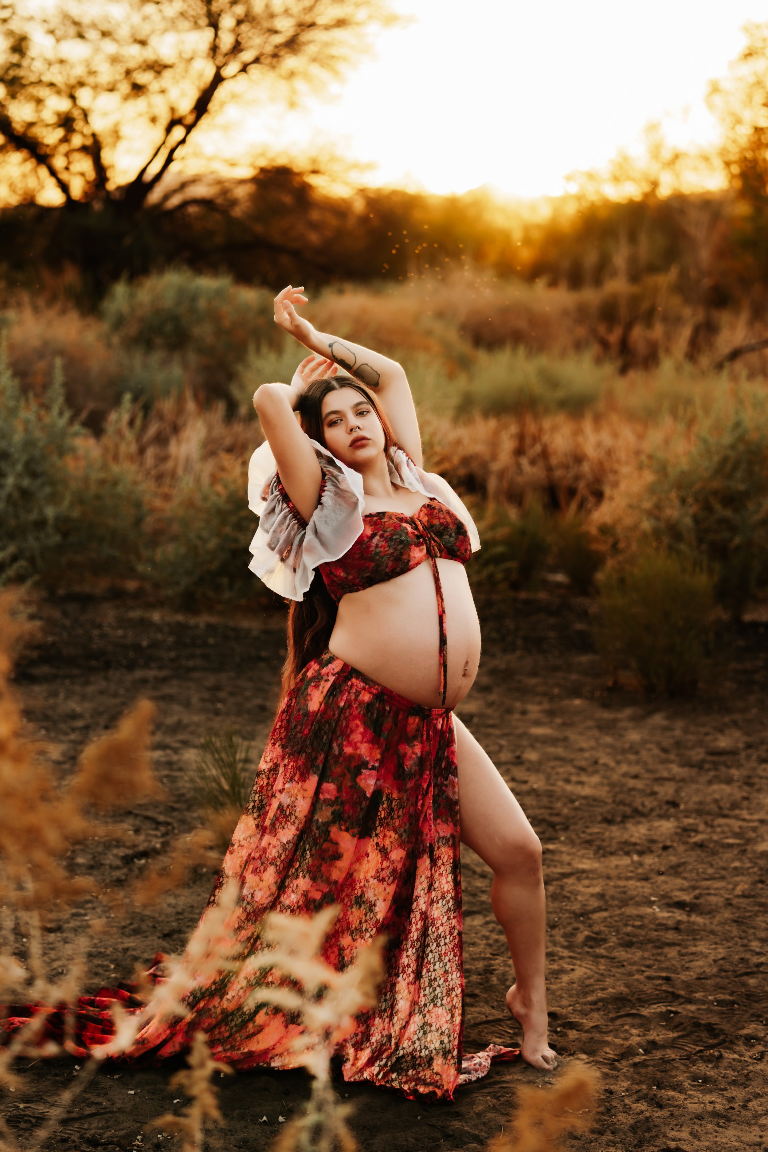 Pregnant woman in a floral dress poses in a desert landscape at sunset, arms raised, surrounded by dry foliage.