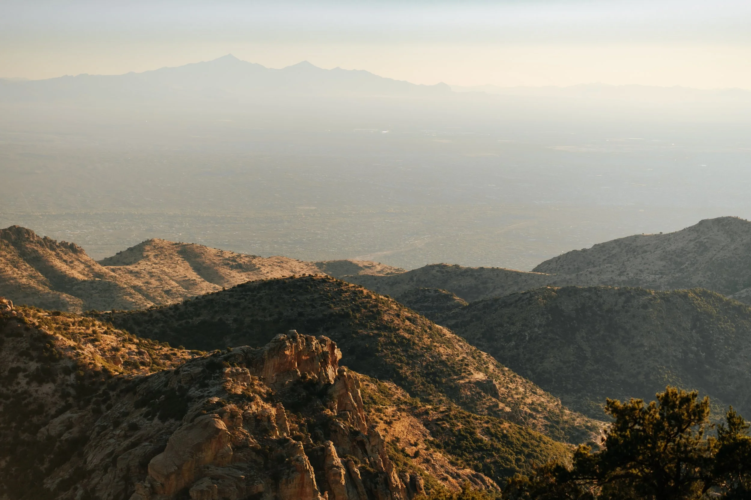 Scenic view of rugged mountains with sparse vegetation, overlooking a hazy valley with distant mountain ranges in the background.