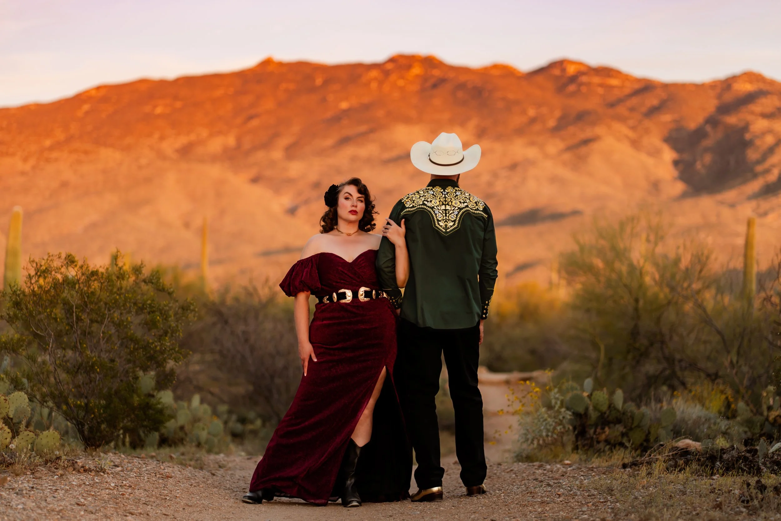 A woman stands facing the camera next to a man facing away from the camera, they both stand in front of an orange mountain at sunset for a couples photoshoot.
