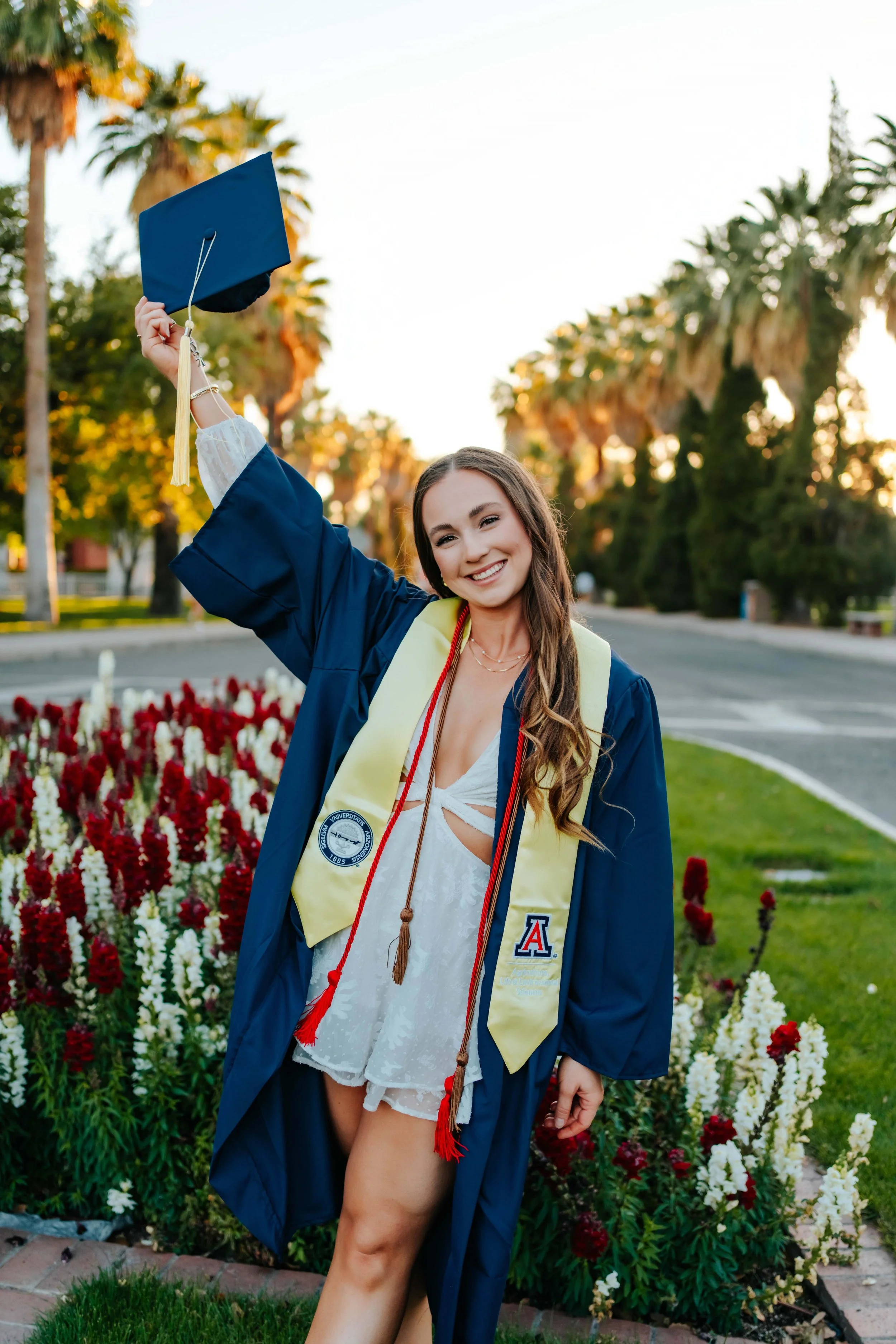 A woman graduating from the University of Arizona smiles and holds her graduation cap in front of flowers and palm trees on the U of A campus at sunset.