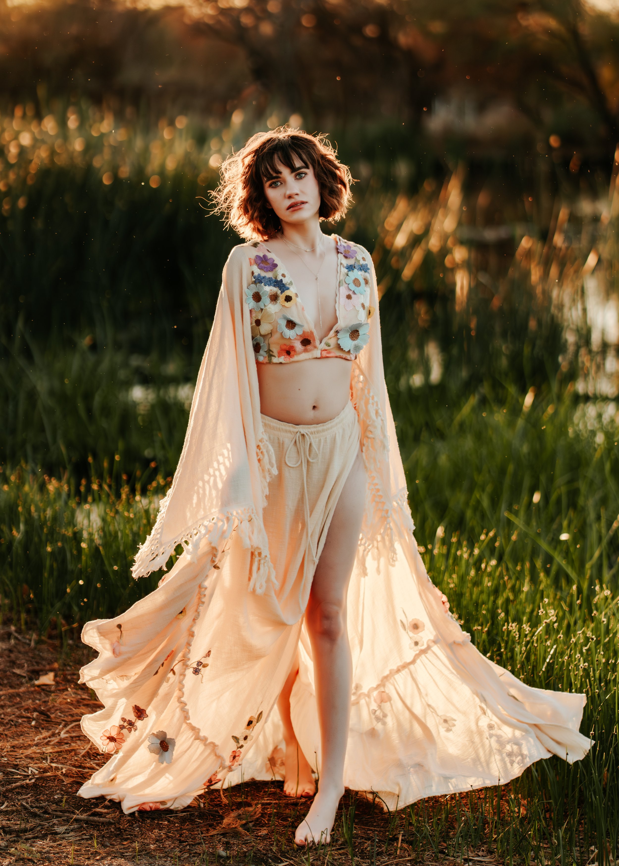 Woman in floral embroidered outfit standing in grassy field at sunset.