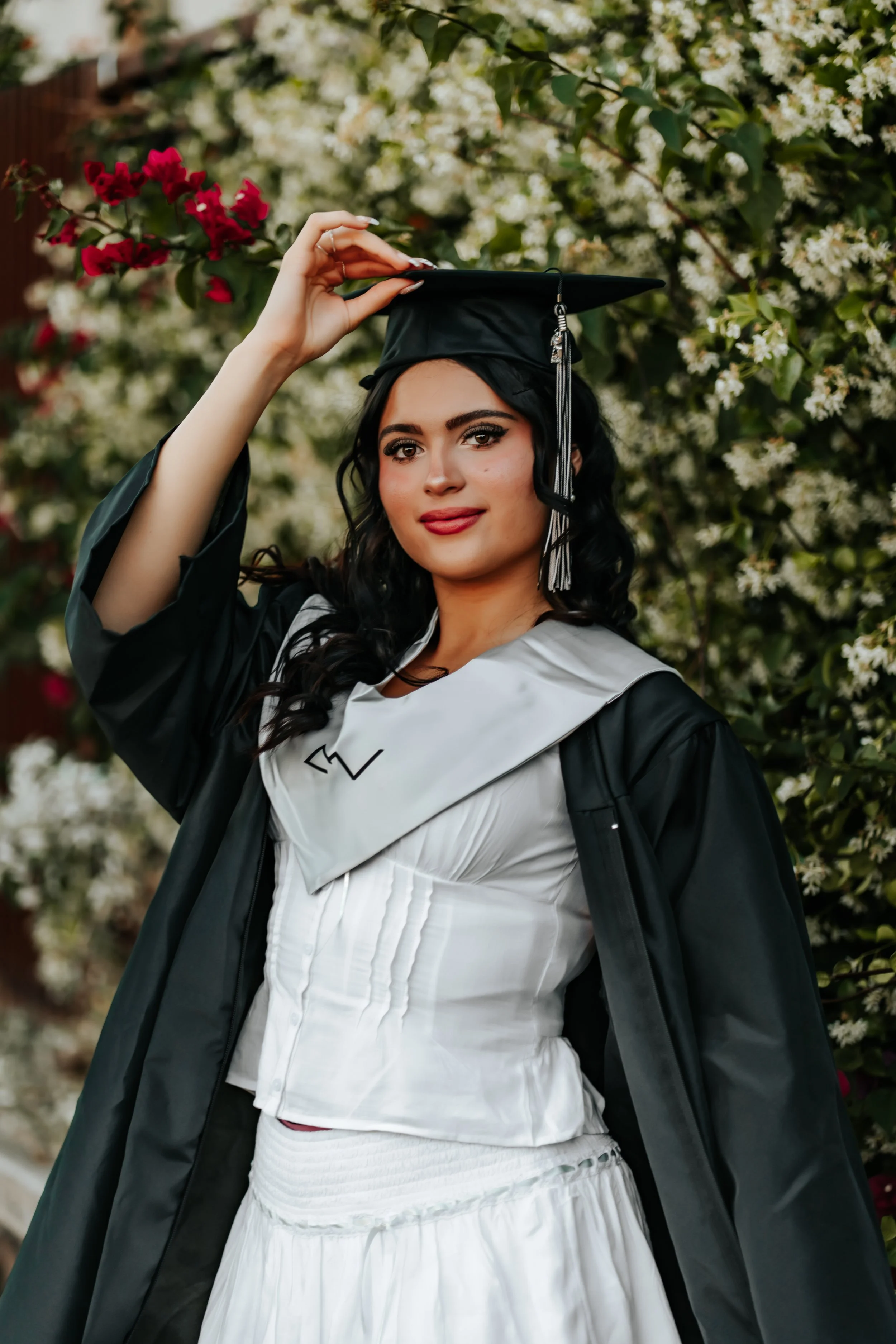 A young woman gazes at the camera in front of a flower wall for her graduation senior photoshoot.