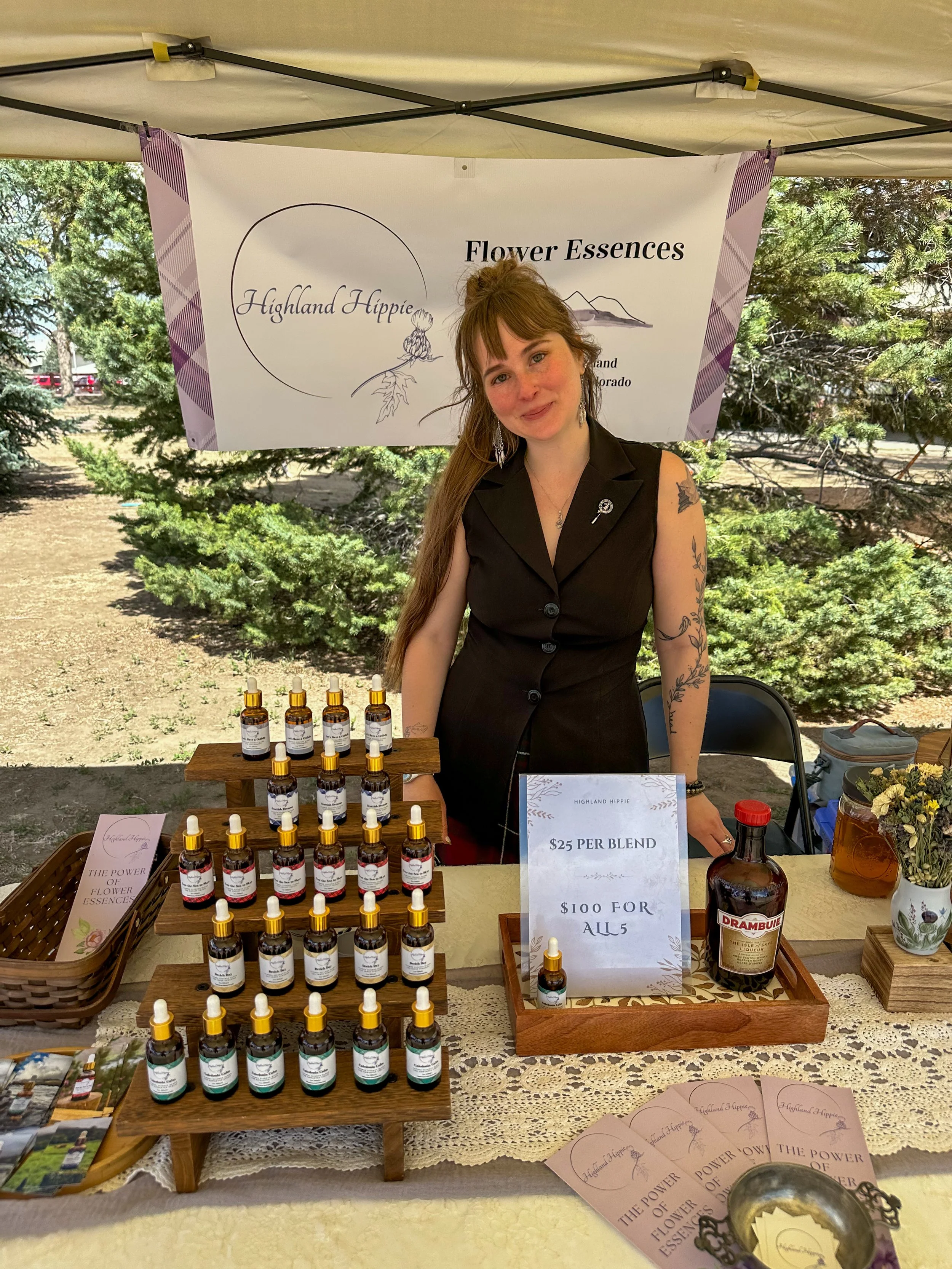 A woman standing behind a table at an outdoor market stall, selling various flower essence bottles. There's a sign indicating the price is $25 per blend or $100 for all five. The backdrop features a banner with the logo 'Highland Hippie' and 'Flower Essences'. The table displays small bottles with droppers, brochures, and a small tray with a plant and a bottle of Drambuie liqueur.
