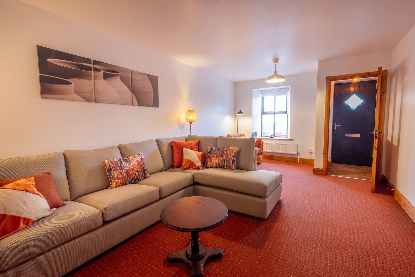 Modern living room with a beige sectional sofa adorned with red and patterned throw pillows, a wooden coffee table, and art on the wall. The room features a red carpet, a floor lamp, a window, and an entry door.