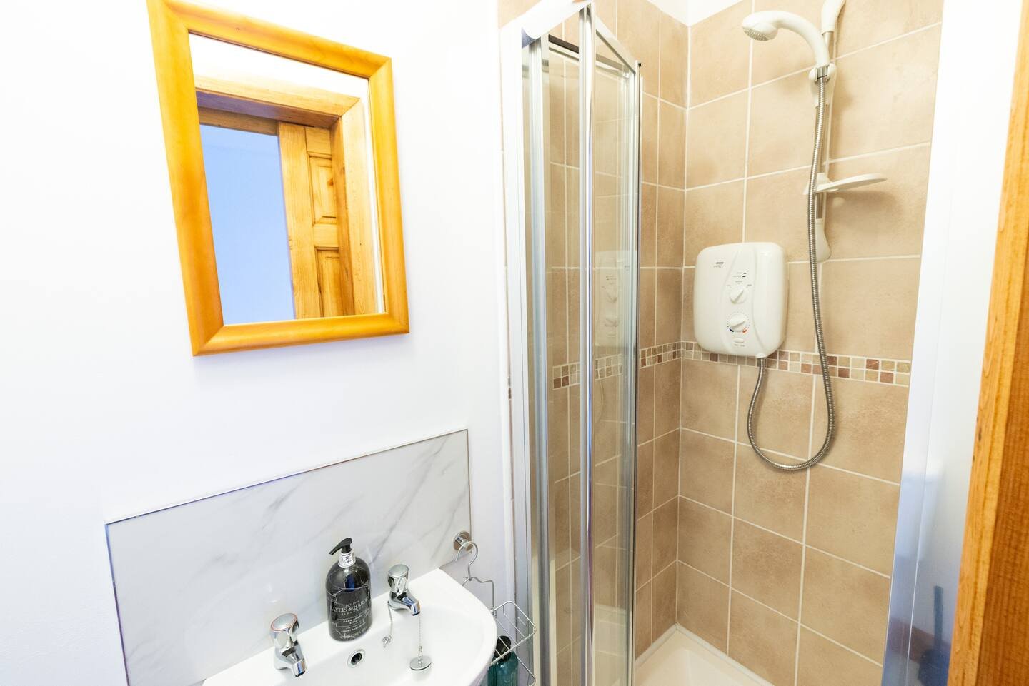 Small bathroom with a corner shower, beige tiles, white sink, mirror with wooden frame, and a wall-mounted electric shower unit.