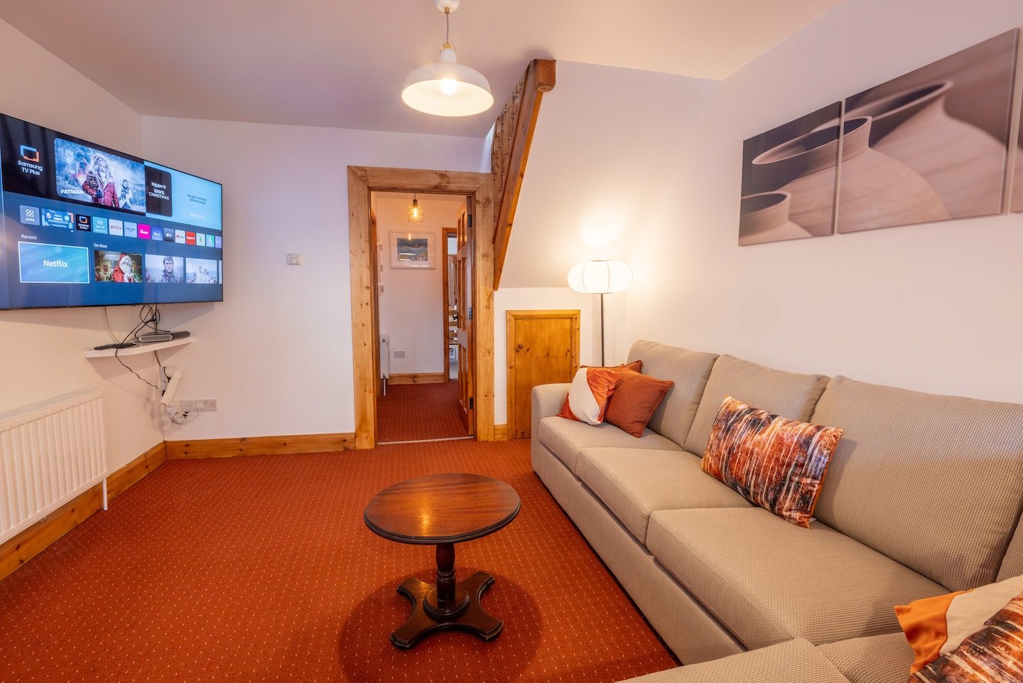 Modern living room interior with a gray sofa, colorful cushions, and a wooden coffee table. A flat-screen TV is mounted on the wall displaying streaming apps. The room has warm lighting, a patterned carpet, and framed artwork above the sofa. There's
