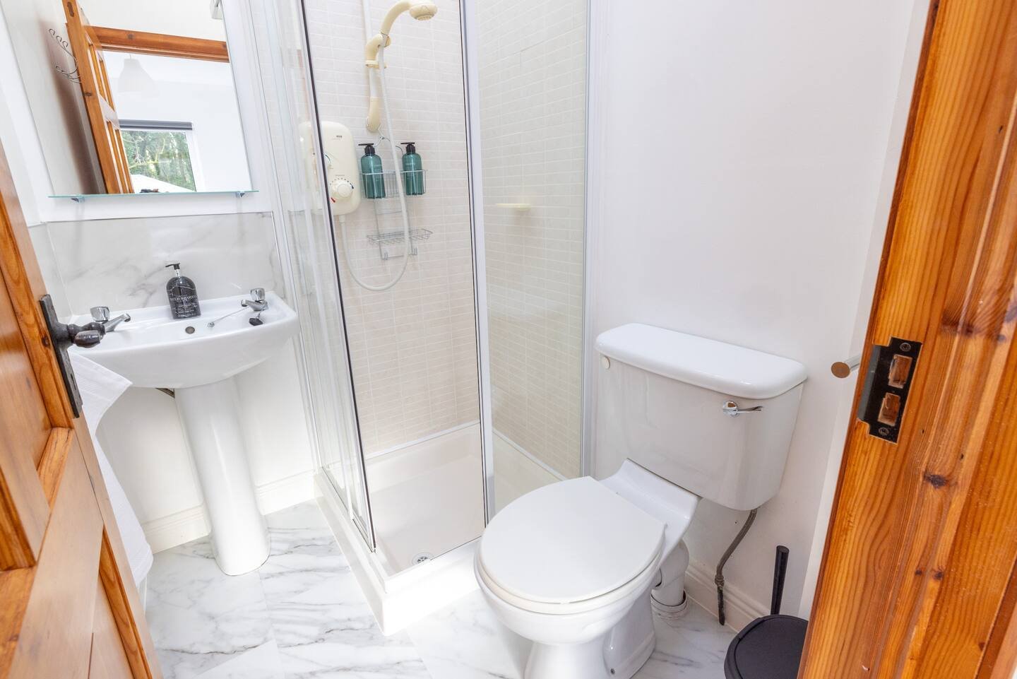 Small bathroom with a shower, toilet, and pedestal sink. The shower has glass doors and is equipped with a handheld showerhead and soap dispensers. The floor is tiled and the room is painted white with wooden accents.