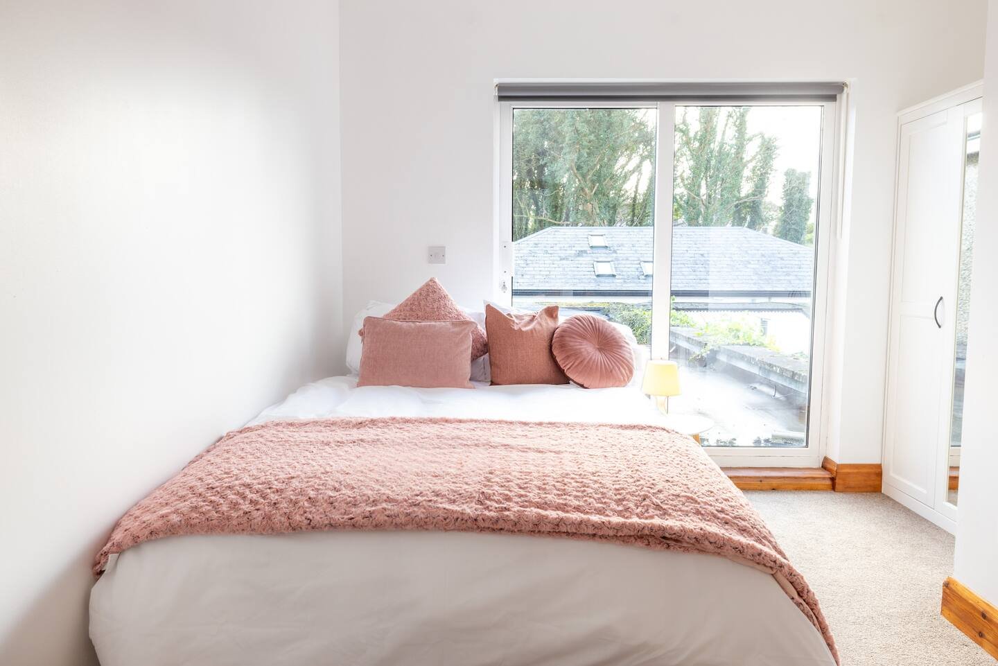 Small bedroom with a white bed, pink pillows, and a pink blanket, next to a large window with an outside view.