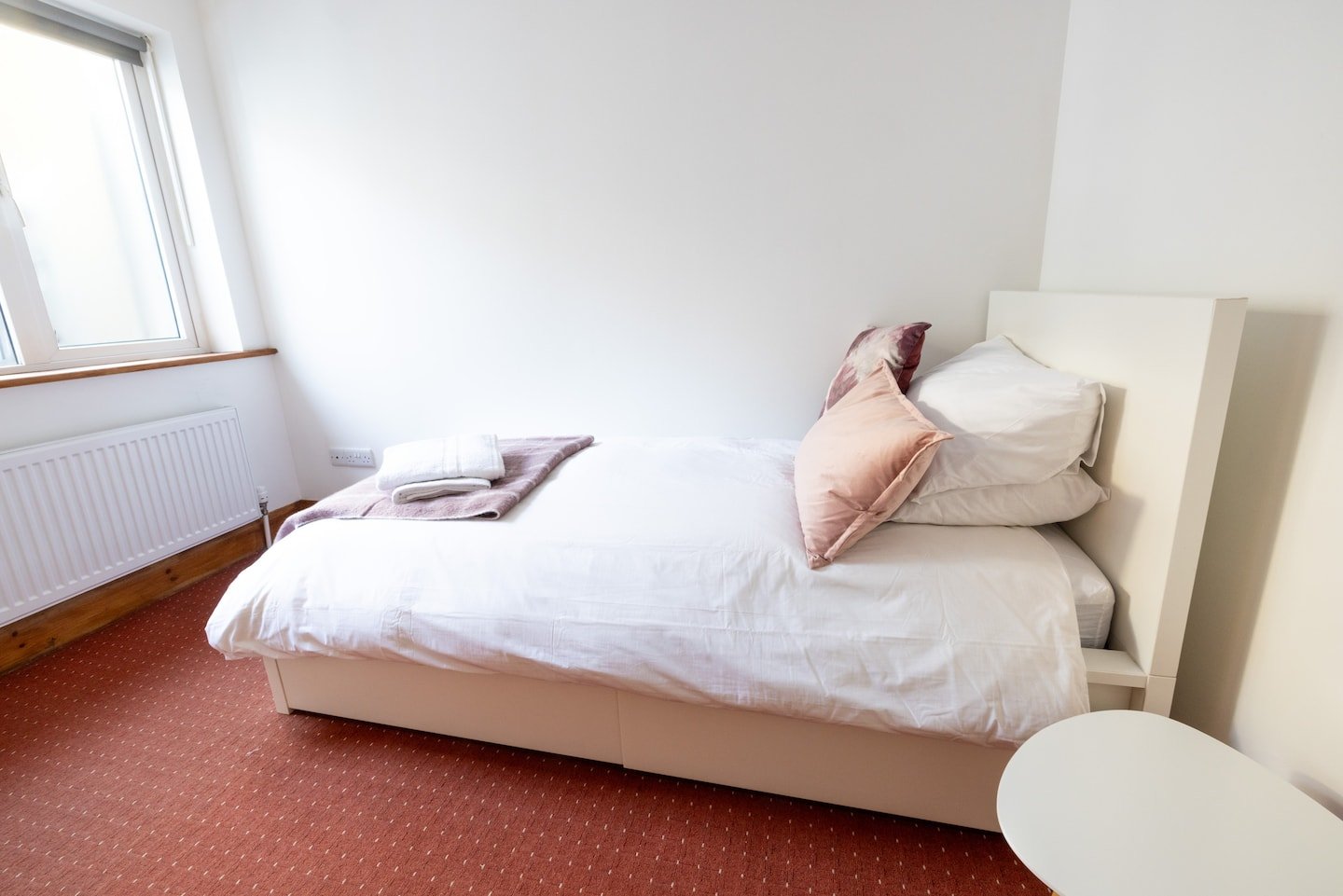 Minimalist bedroom with a single bed, white bedding, pink and white pillows, a side table, and a window.