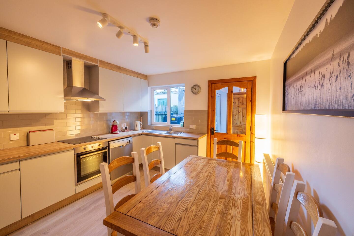 Modern kitchen with wooden dining table, chairs, light cabinets, stainless steel appliances, and tile backsplash.
