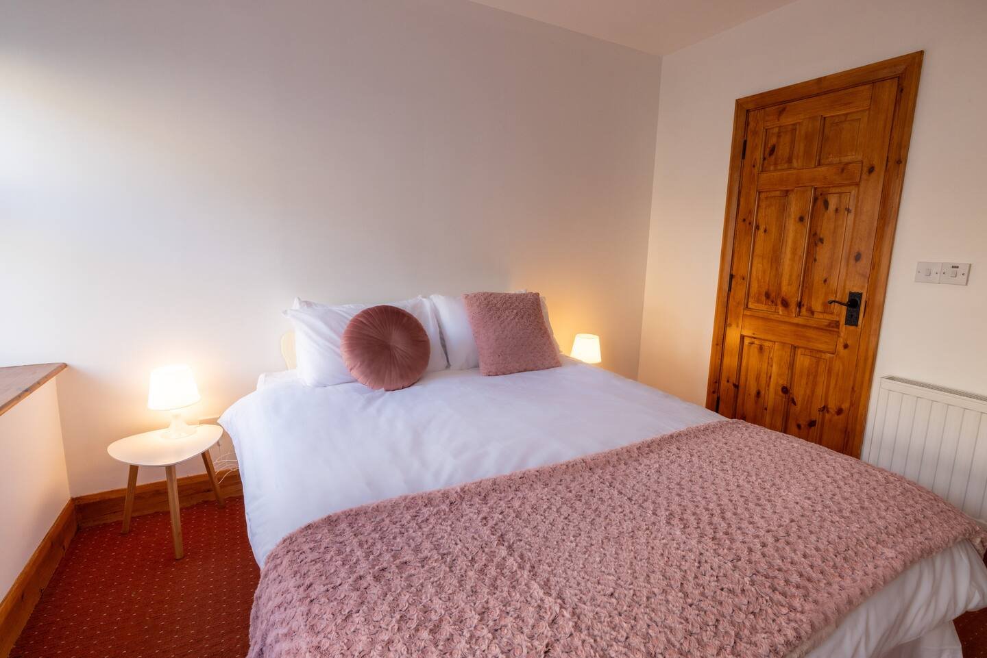 Cozy bedroom with a white bed, pink decorative pillows, and a knitted blanket. Wooden door and small bedside tables with lamps.
