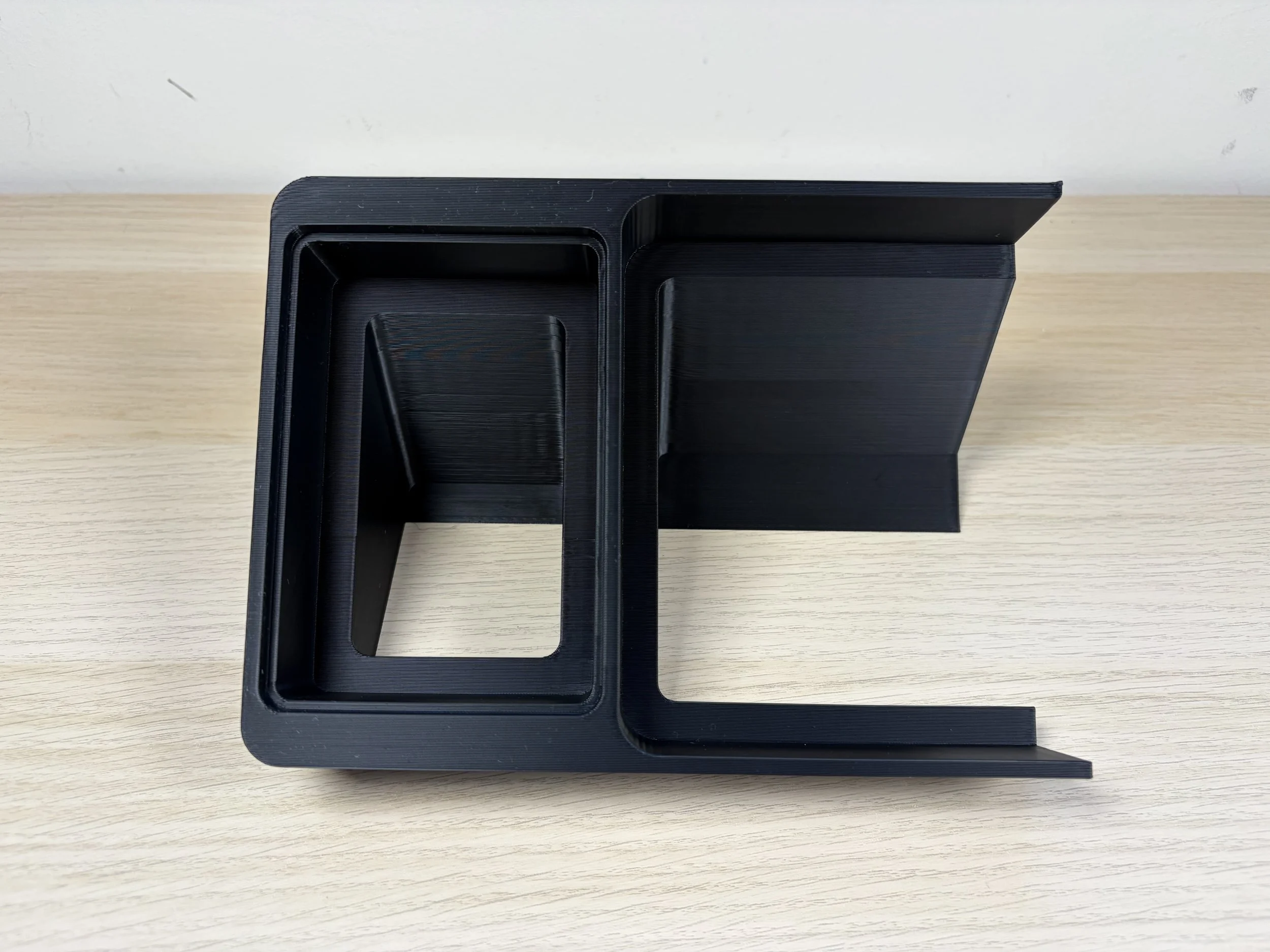 3D-printed black plastic object with two open square compartments, placed on a light-colored wooden surface.
