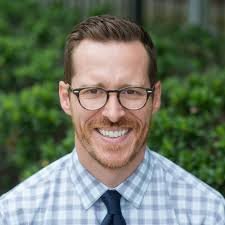 A man with glasses and a beard smiling outdoors with greenery in the background.