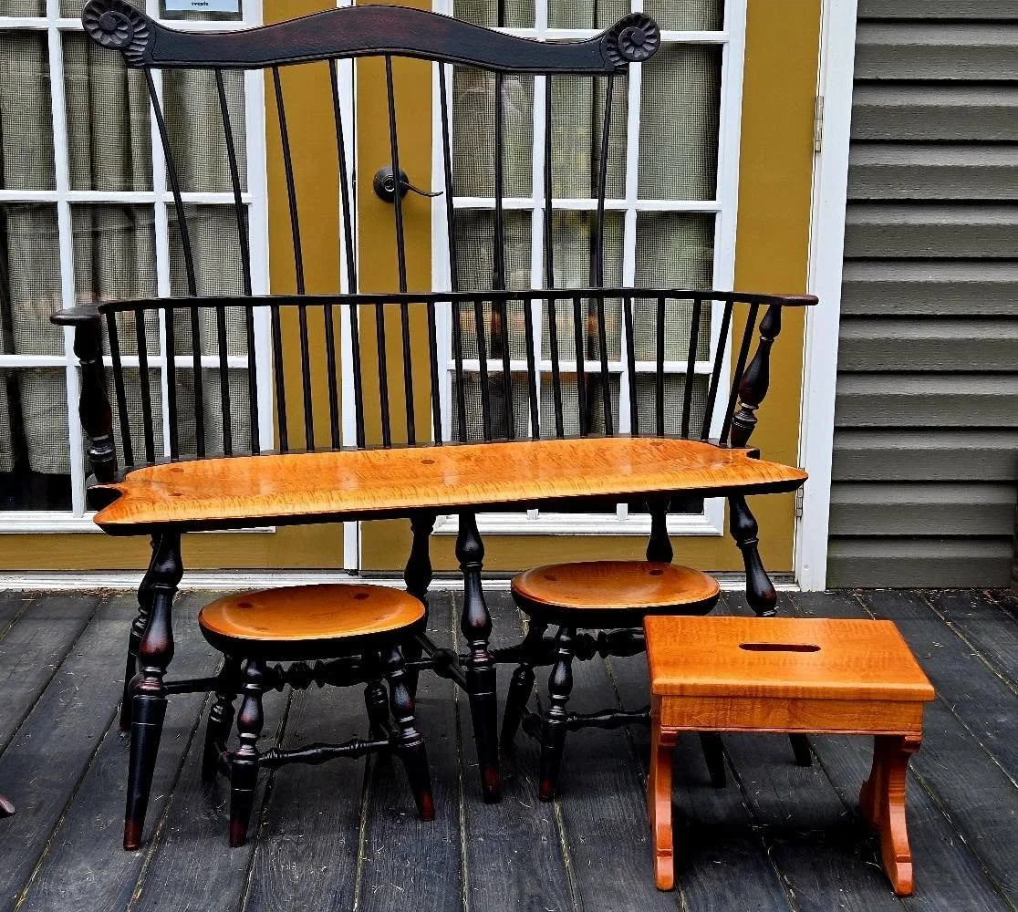 A wooden swing with a backrest, two stools, and a small wooden table on a dark wooden porch. The swing and stools are made of dark and light wood, in front of a glass door with a yellow frame and gray siding.