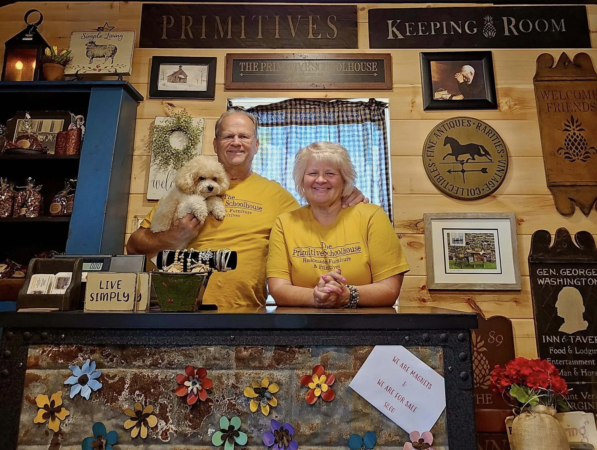 Two smiling people in yellow T-shirts standing behind a counter with a small white dog. The wall behind them is decorated with signs, framed pictures, and wooden ornaments. The counter has colorful flower decorations and a sign that says 'WE ARE MAGNETS & WE ARE FOR SALE $8.00'.