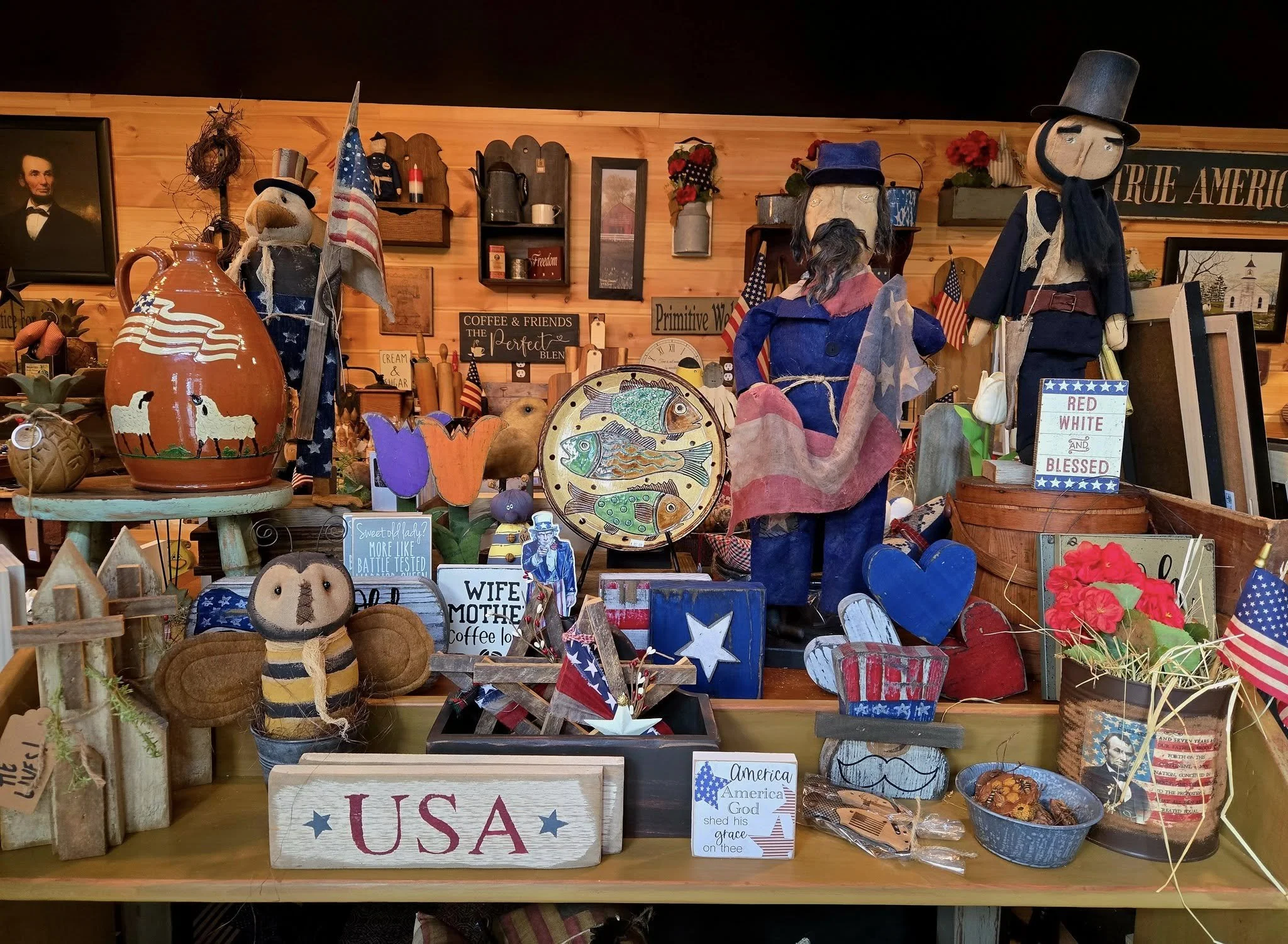 A display of patriotic and Americana-themed decorations, including stuffed dolls dressed in Union and Confederate uniforms, American flags, a painted jug with sheep, wooden hearts, flowers, and various signs with patriotic messages, set against a wooden wall background.