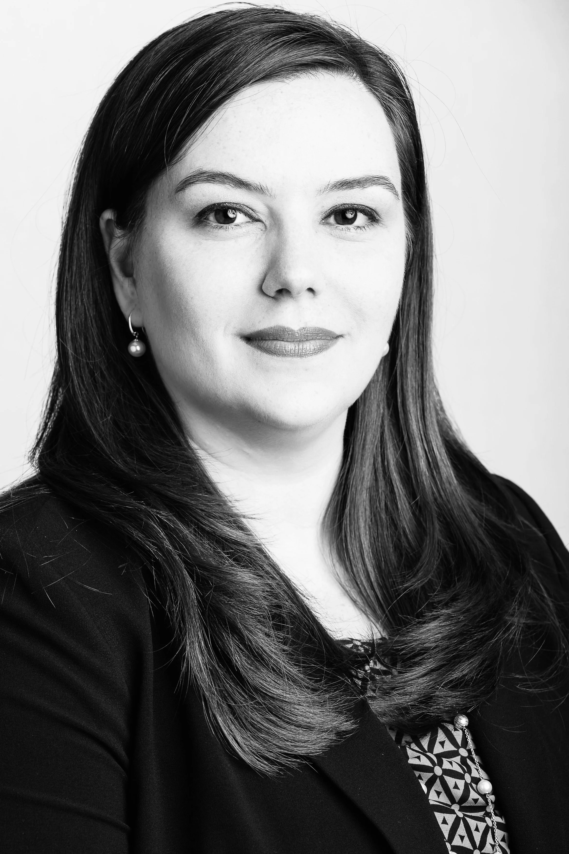 Black-and-white portrait of a woman with dark hair, wearing a patterned top, pearl earrings, and a blazer, looking at the camera