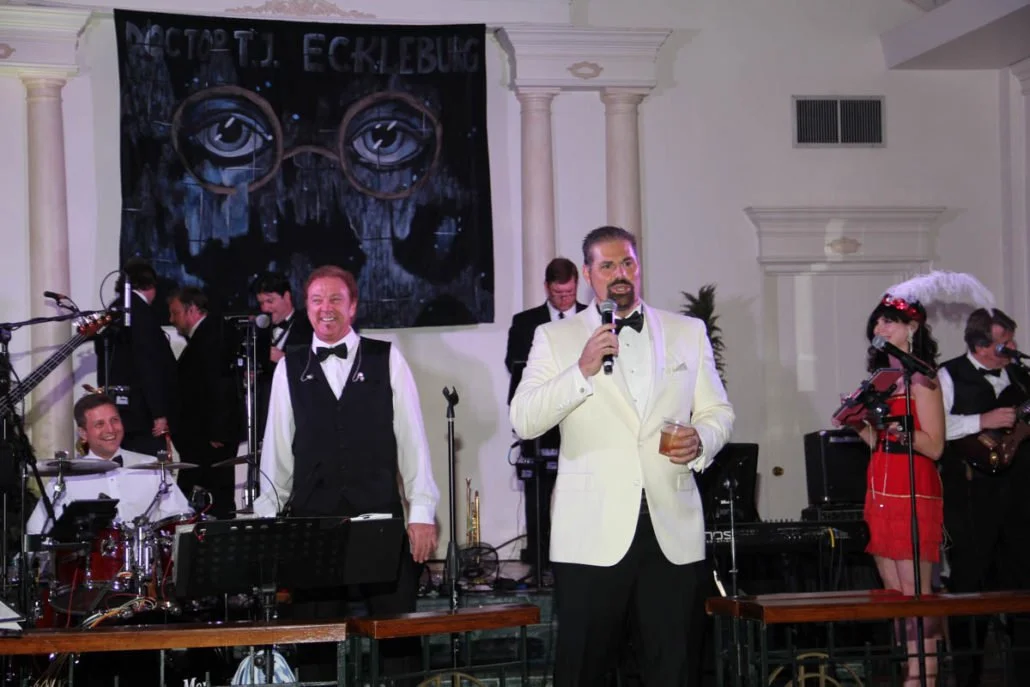 Band performing on stage with a man in a white tuxedo holding a microphone and drink, a woman in a red dress with a feathered headpiece singing, and a large banner with eye artwork in the background.