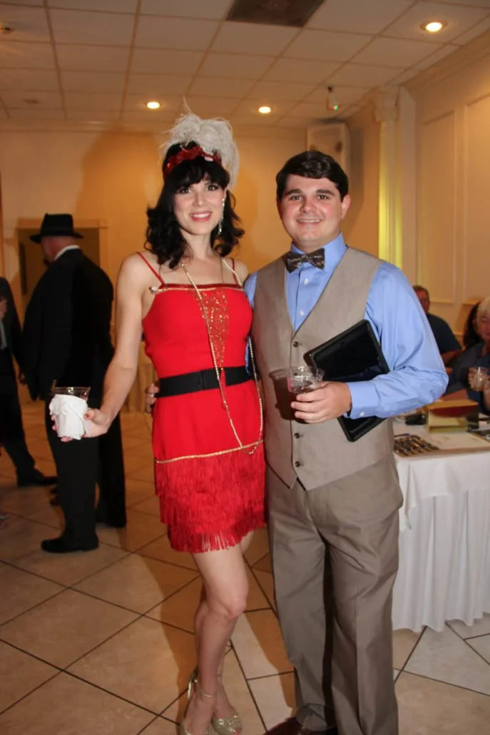 Two people dressed in vintage-inspired clothing at a party. The woman is wearing a red dress with fringe, a feather headpiece, and heels. The man is wearing a blue shirt, brown vest, bow tie, and slacks, holding a drink and a tablet.