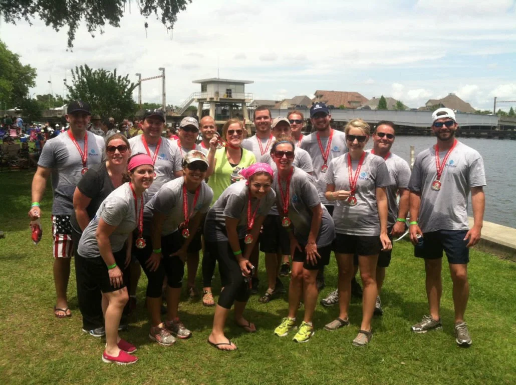 A group of people wearing matching gray shirts and medals, posing outdoors near a waterway on a grassy area, after participating in a running or outdoor event.