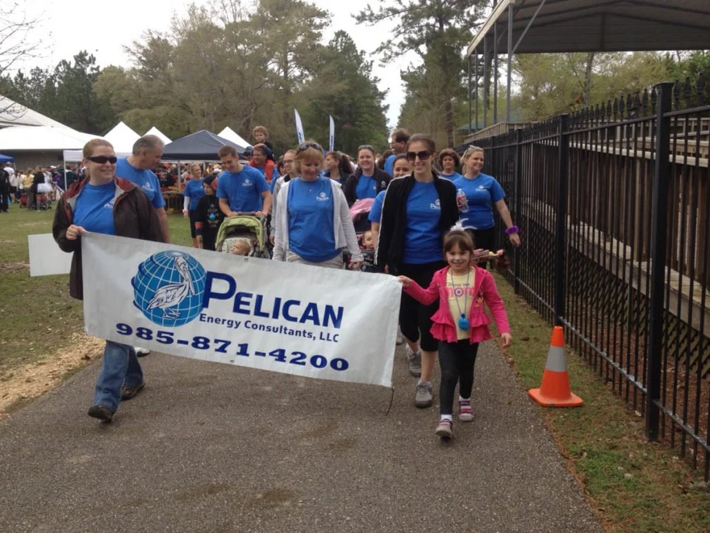 A group of people marching in an outdoor event, holding a banner for Pelican Energy Consultants, LLC, with a young girl at the front smiling and holding the hand of a woman.