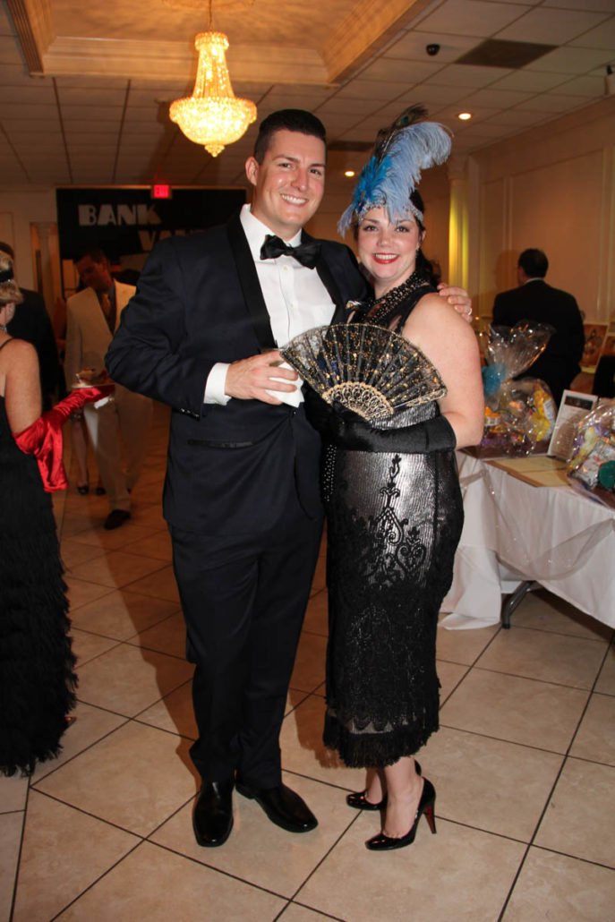 A man in a tuxedo with a bow tie and a woman in a vintage-style black dress with lace and sequin details, holding a decorative fan. She is wearing a feathered blue headpiece, and they are posing at a formal event or party in a banquet hall.