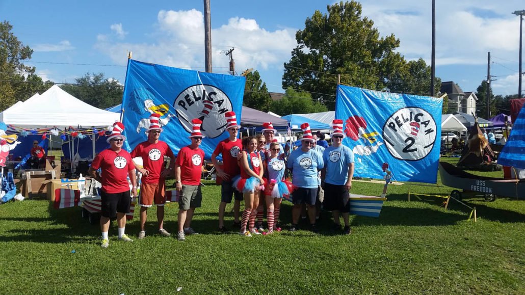 Group of people dressed in red and blue shirts and striped hats, standing in front of Pelican-themed banners and decorations at an outdoor event.